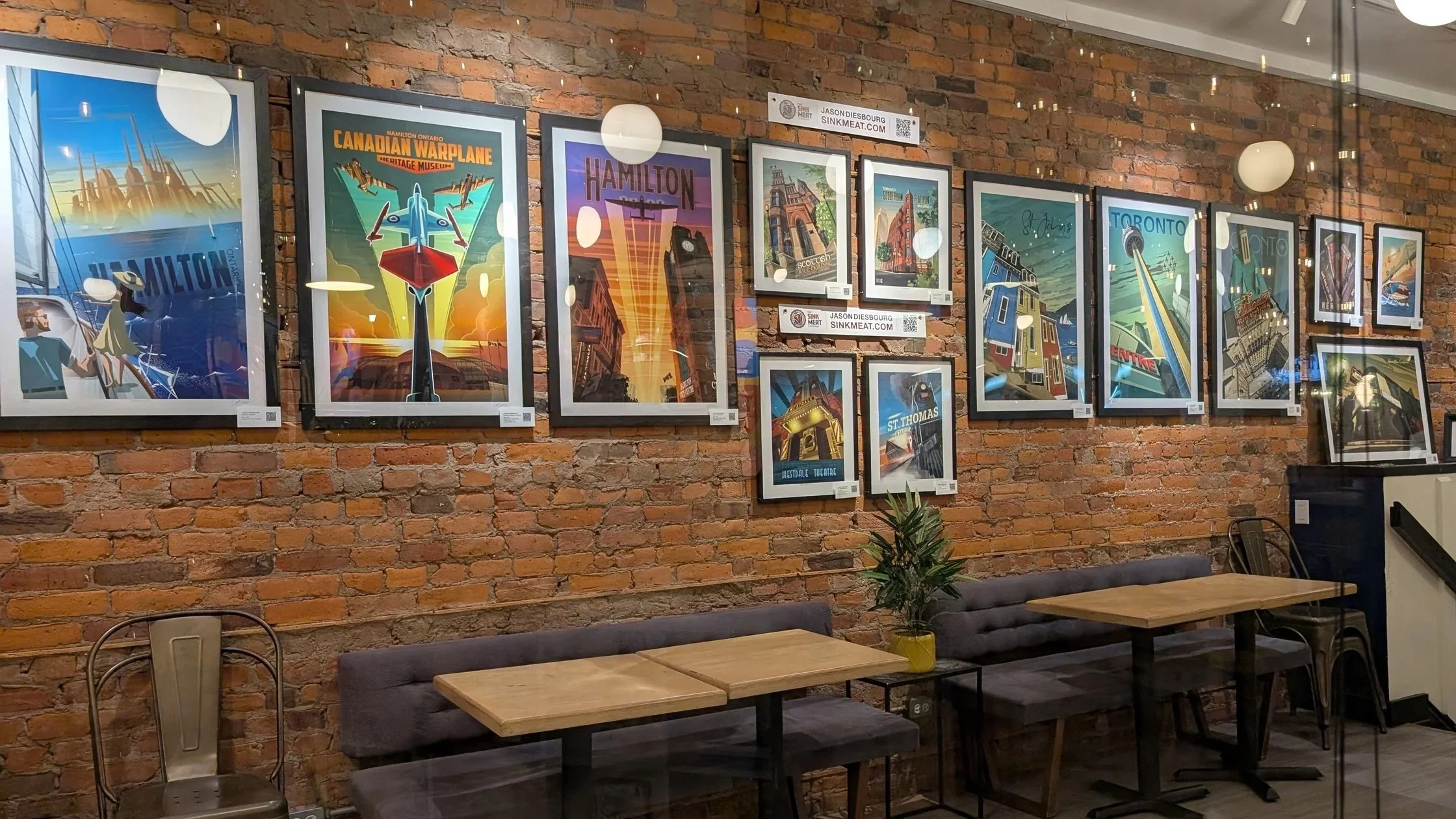 Series of framed posters of iconic Toronto landmarks and scenes on a brick wall in a cafe or gallery, with a small table and chairs in the foreground.