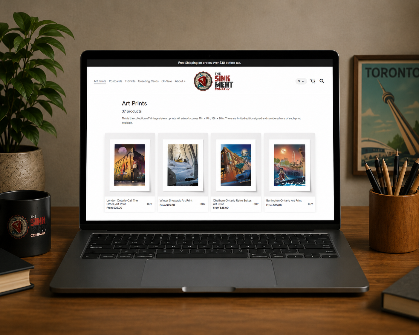 Laptop on a wooden desk displaying an art prints online store, with potted plants and books on either side, and a Toronto skyline picture on the wall behind.