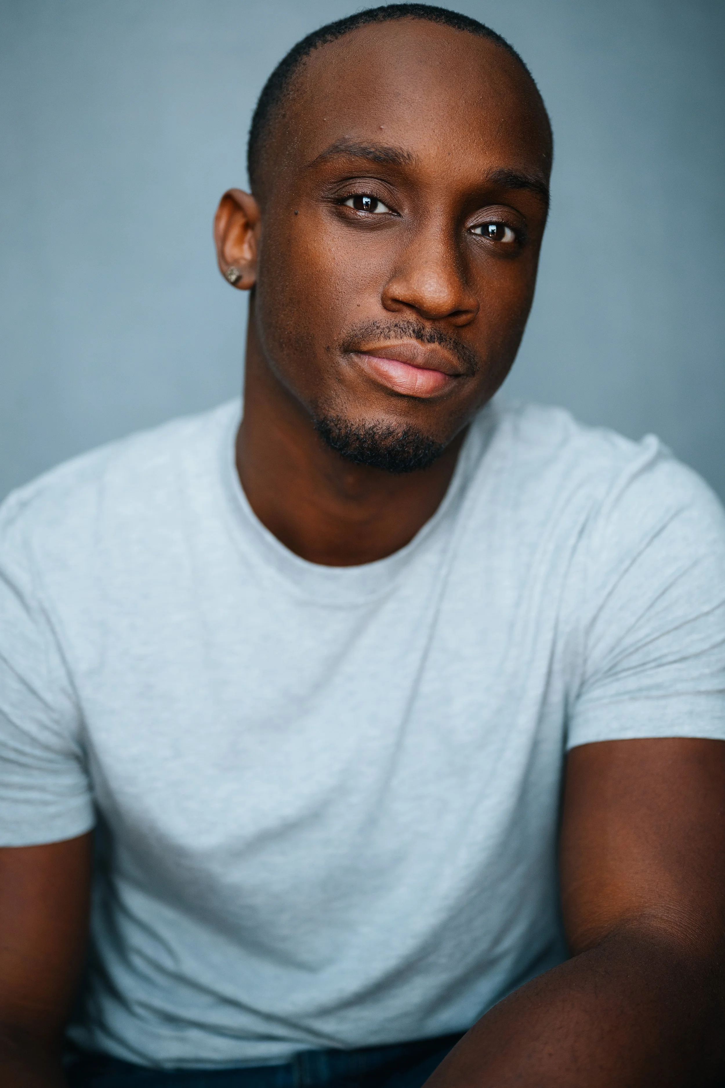 Portrait of a young man with a close-cropped haircut, dark skin, and a slight smile, wearing a light gray t-shirt, against a plain grayish background.