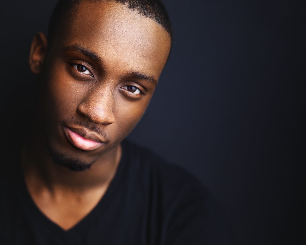 Close-up portrait of a young man with short black hair, light beard, and a slight smile, wearing a black shirt against a dark background.