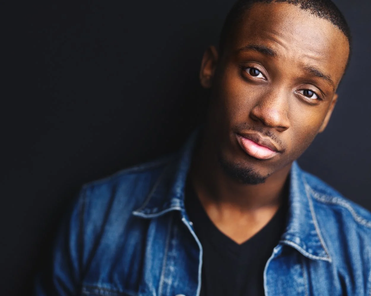 Close-up portrait of a young man with short hair, wearing a denim jacket over a black shirt, against a solid black background.
