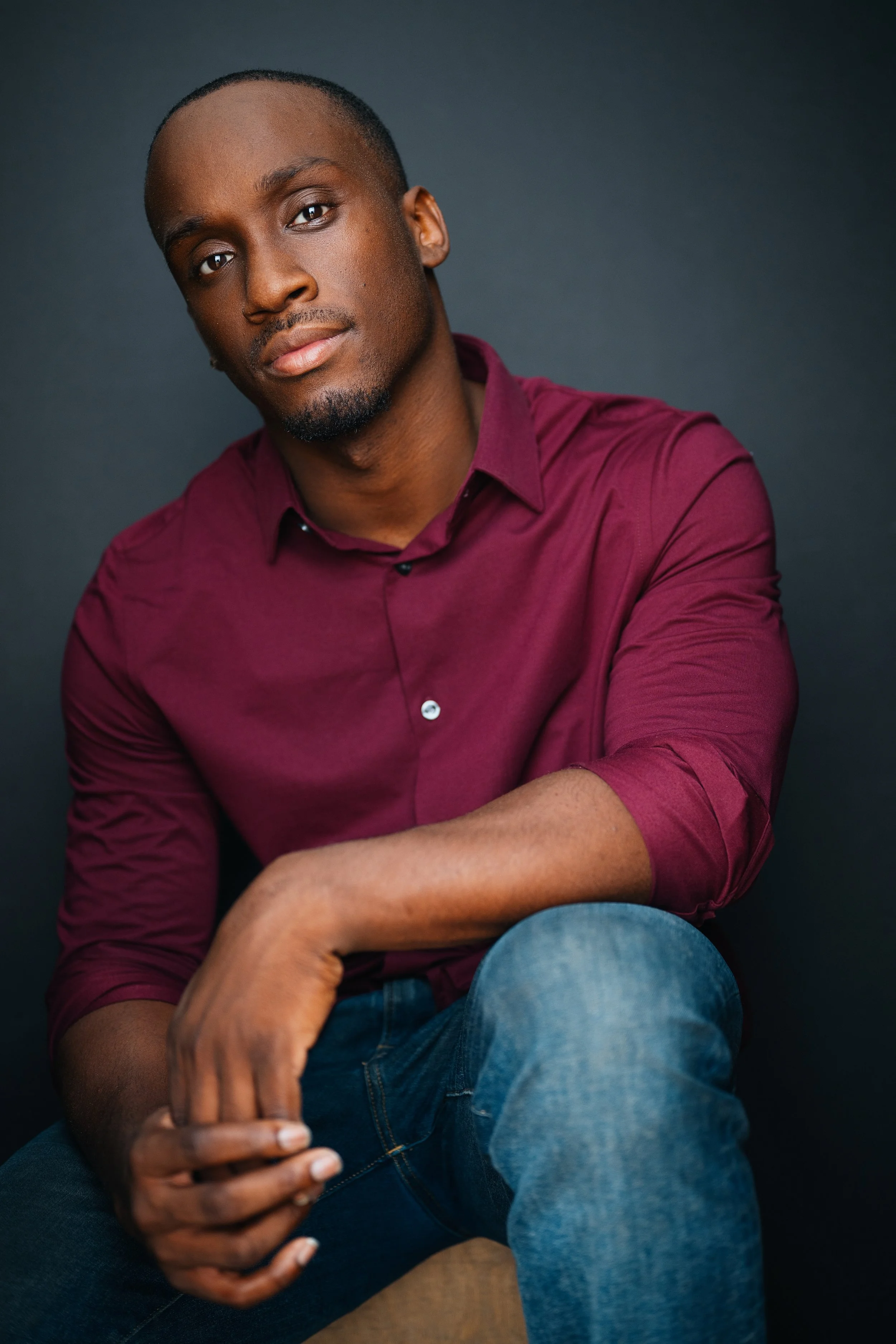 A young Black man with a short haircut and goatee sitting against a dark background, wearing a maroon button-down shirt and jeans, looking directly at the camera with a relaxed expression.