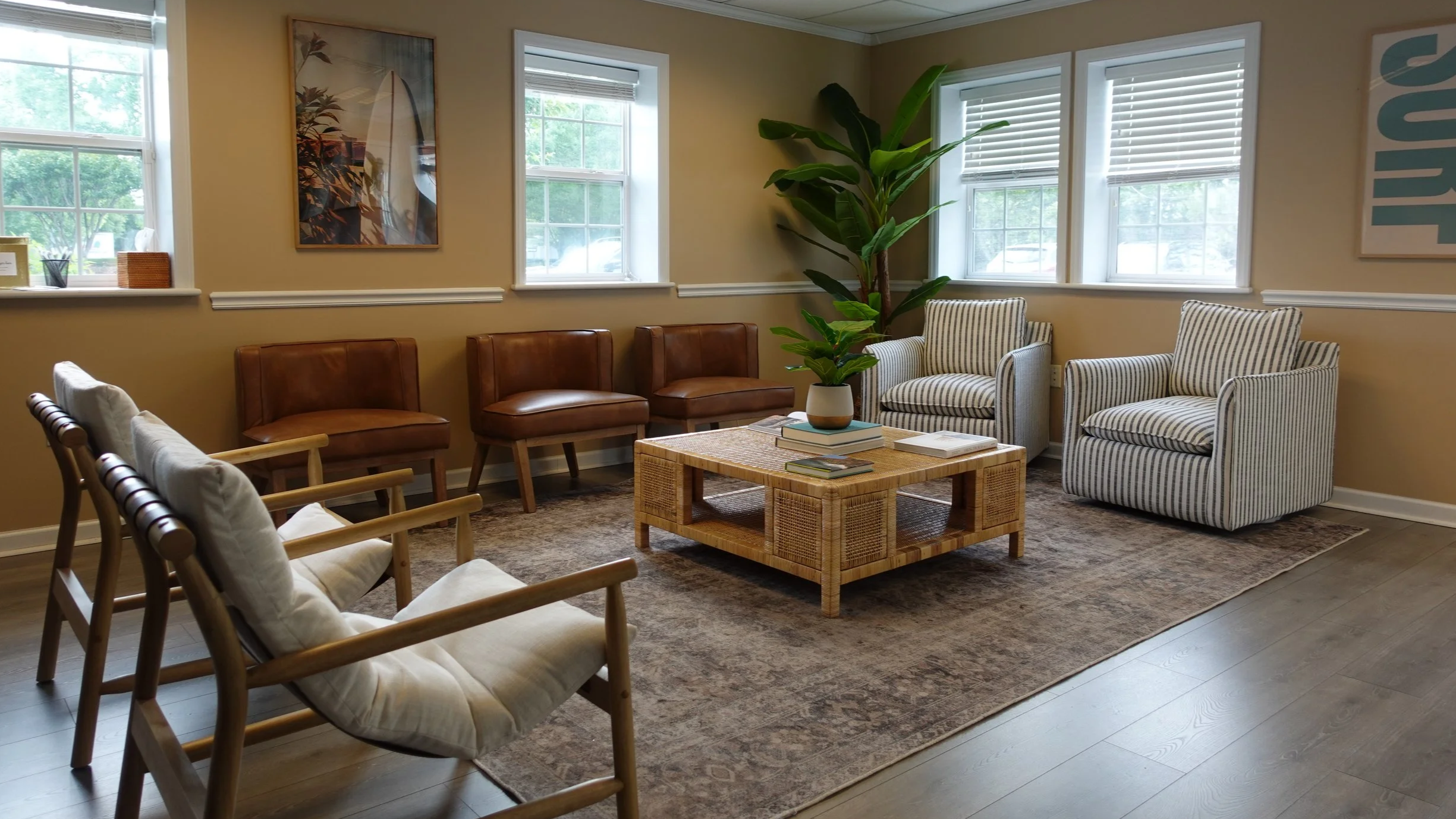 A cozy waiting room with beige walls, multiple windows letting in natural light, and various chairs including wooden armchairs with white cushions, leather chairs, and striped upholstered armchairs. A wooden coffee table with books and a potted plant is in the center. There is a large plant near the window, and framed artwork on the wall.