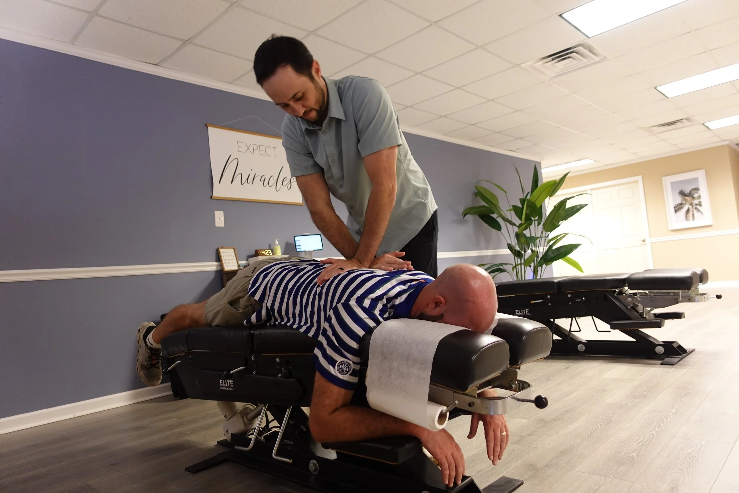 A chiropractor is performing a spinal adjustment on a male patient lying face down on an adjustable chiropractic table in a treatment room. The room has a purple accent wall with a sign that reads 'Expect Miracles', a plant, and framed artwork of a palm tree.