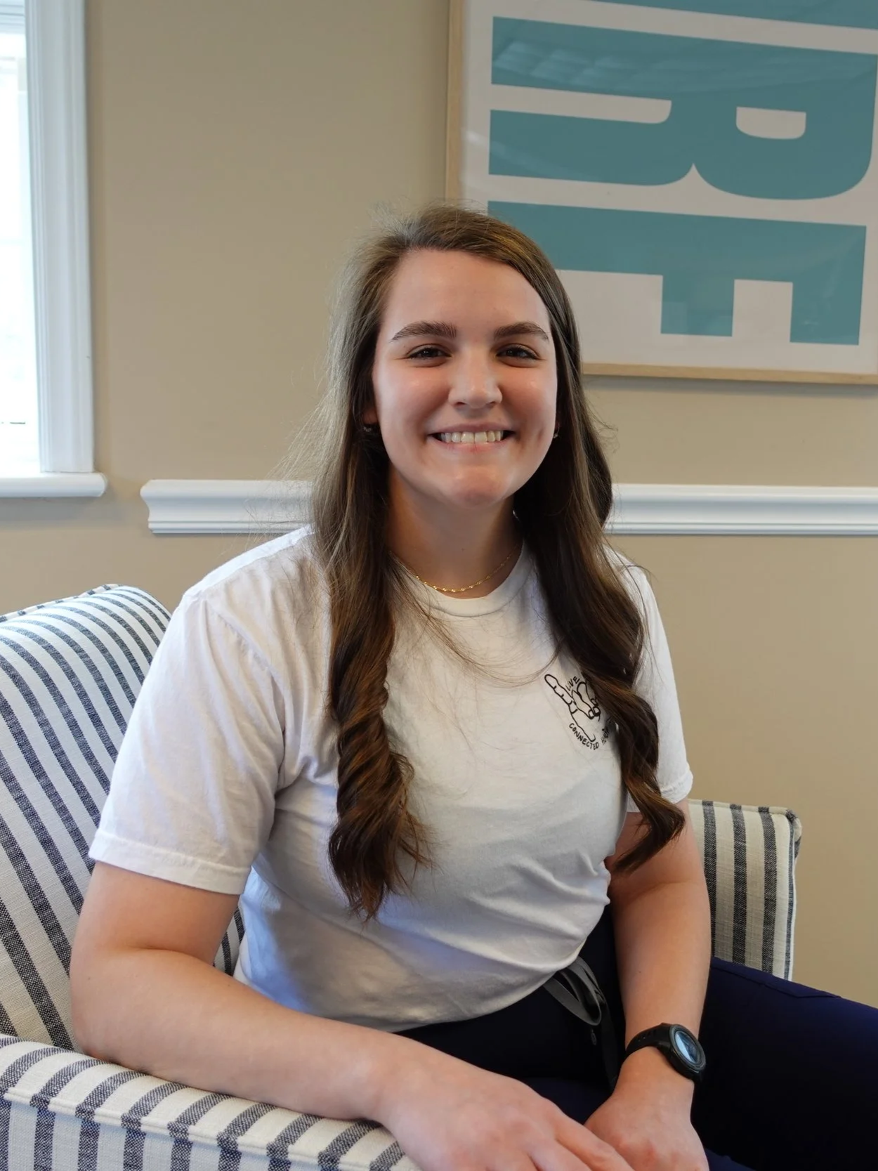 A young woman with long brown hair sitting on a striped armchair, smiling at the camera. She is wearing a white T-shirt, black pants, and a black wristwatch. Behind her is a beige wall with a large blue and white decorative display.