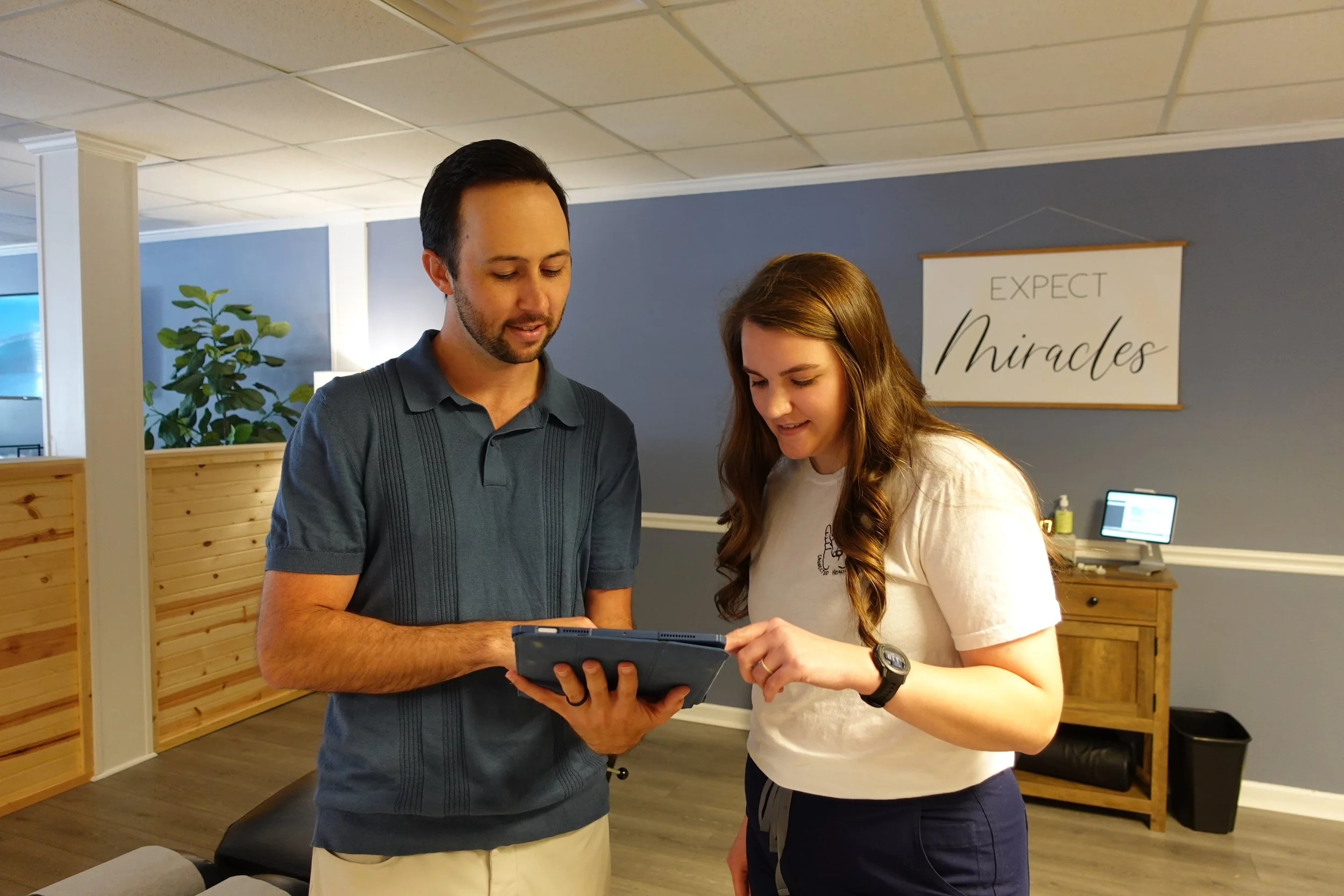 A male chiropractor and female chiropractor assistant, are reviewing patient nervous system scans on a tablet together in an indoor setting with a sign in the background that reads "EXPECT Miracles."