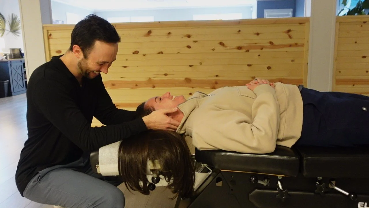 A chiropractor adjusting a woman's neck while she is lying on a chiropractic table, smiling in a clinic with wooden dividers.
