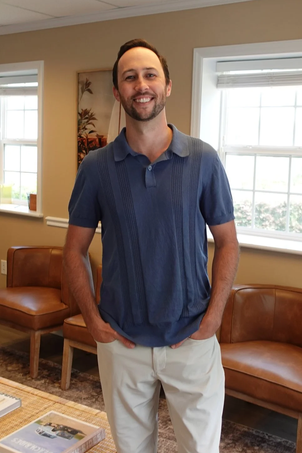 A man with a beard and mustache smiling while standing in a room with beige walls and windows, wearing a blue polo shirt and light-colored pants.
