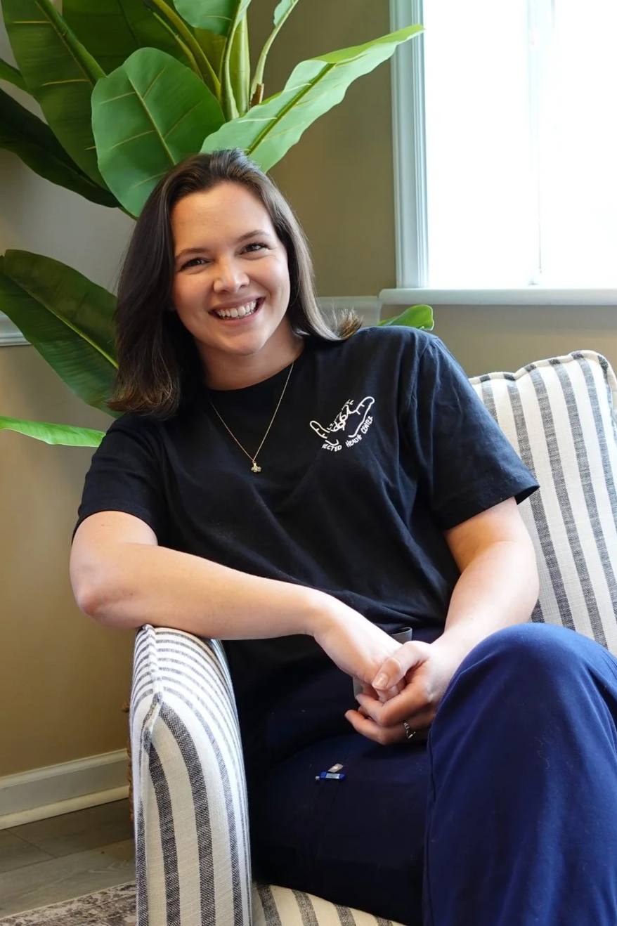 A young woman with shoulder-length brown hair sitting on a striped armchair, smiling, wearing a black t-shirt and a delicate necklace, with large green leaves of a plant behind her near a window.