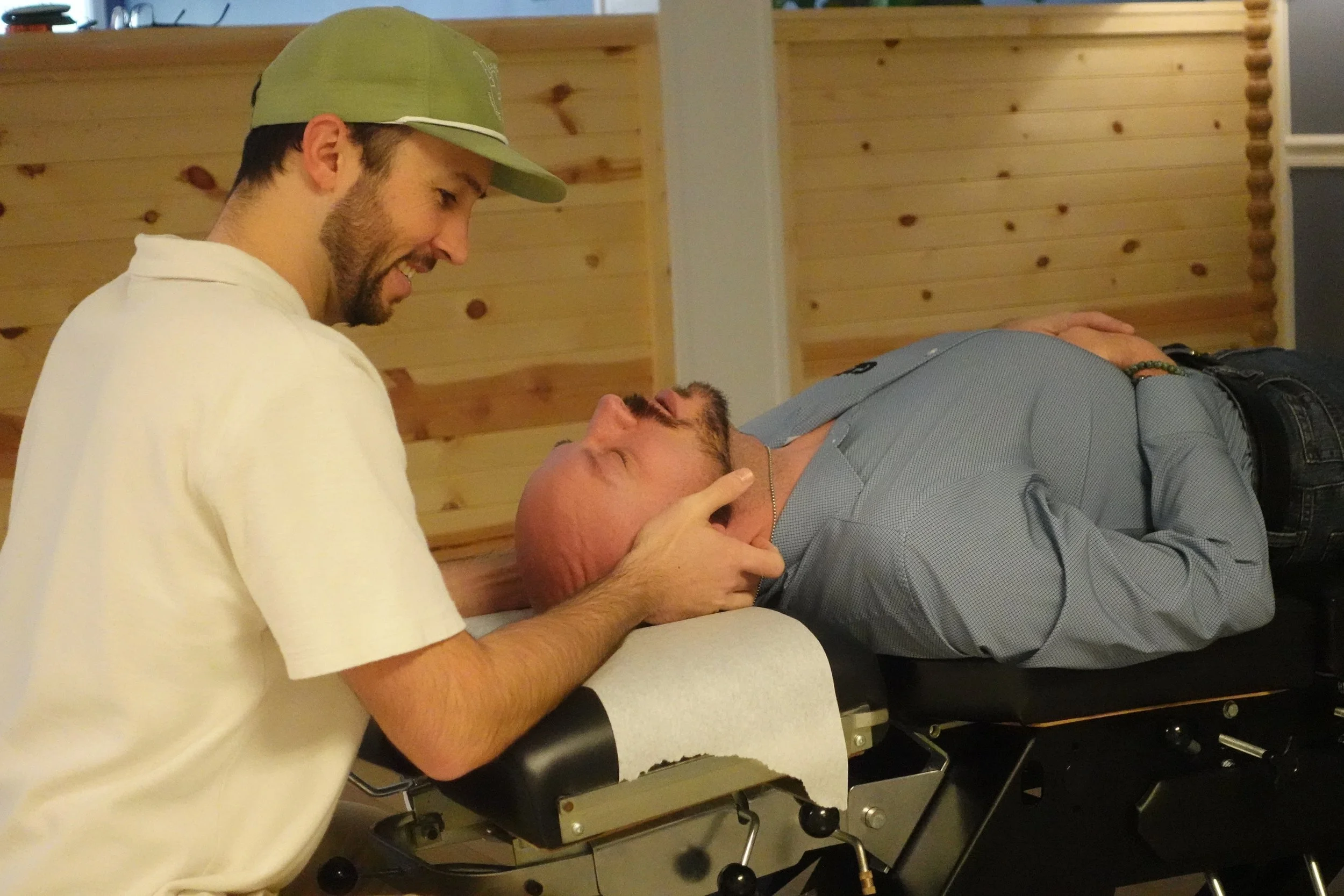 A man lying on a medical examination table, with his head supported by a woman who is holding his face with both hands. The woman is smiling and looking at the man. The background features wooden wall paneling.
