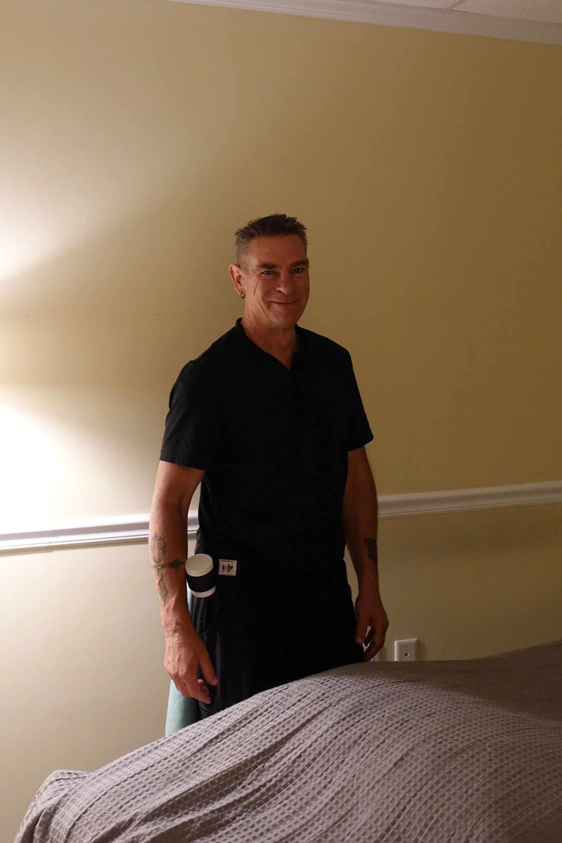 A middle-aged man with short dark hair and tattoos on his right arm, wearing a black shirt, standing in a room with yellow walls, smiling at the camera.