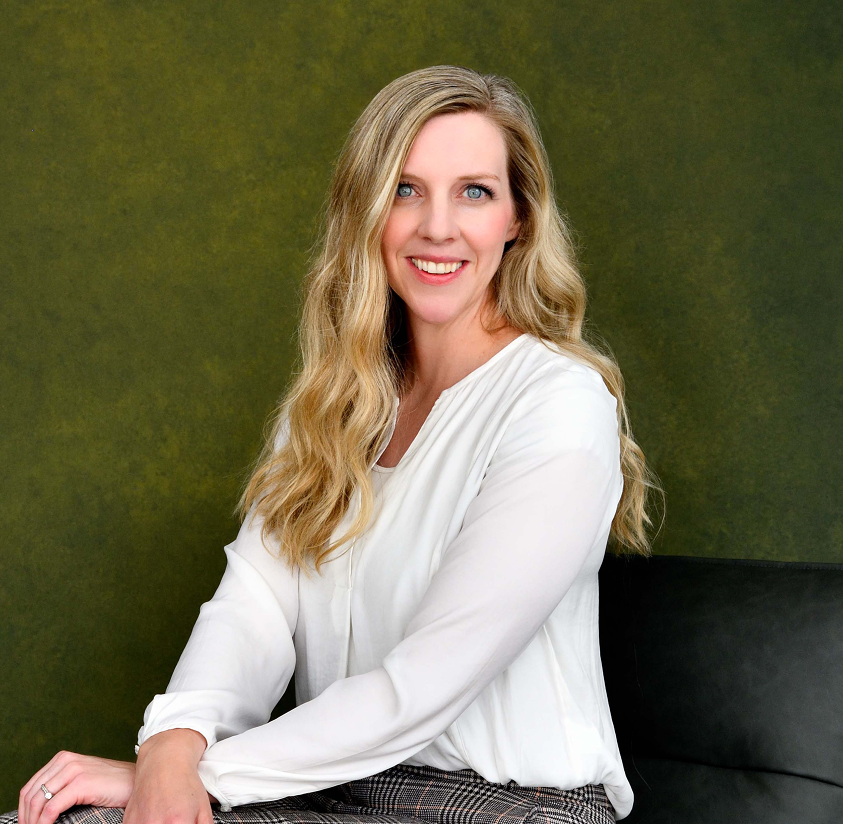 A woman with long wavy blonde hair and blue eyes smiling, wearing a white blouse and checkered pants, seated against an olive green background.