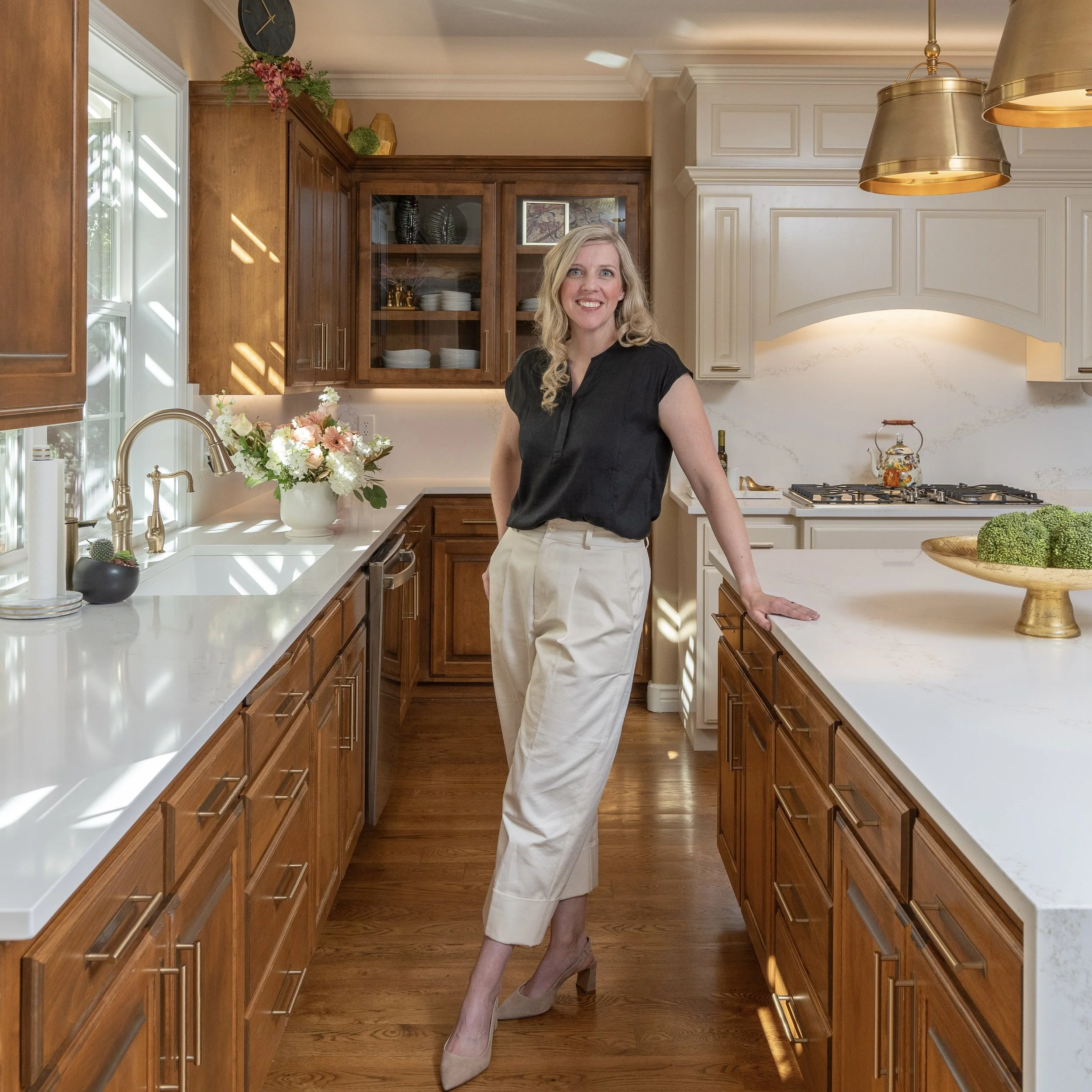 A woman with blonde curly hair smiling, wearing a black top and beige pants, standing in a spacious kitchen with wooden cabinets, white countertops, a window with sunlight streaming in, and decorative flowers on the countertop.