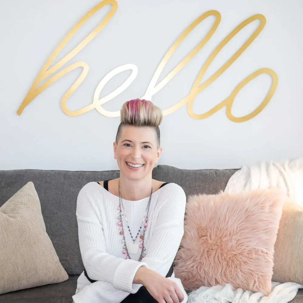 A woman with short, styled hair with pink highlights sitting on a gray couch, smiling at the camera, in front of a white wall with a large gold cursive "hello" sign, surrounded by beige and pink pillows.