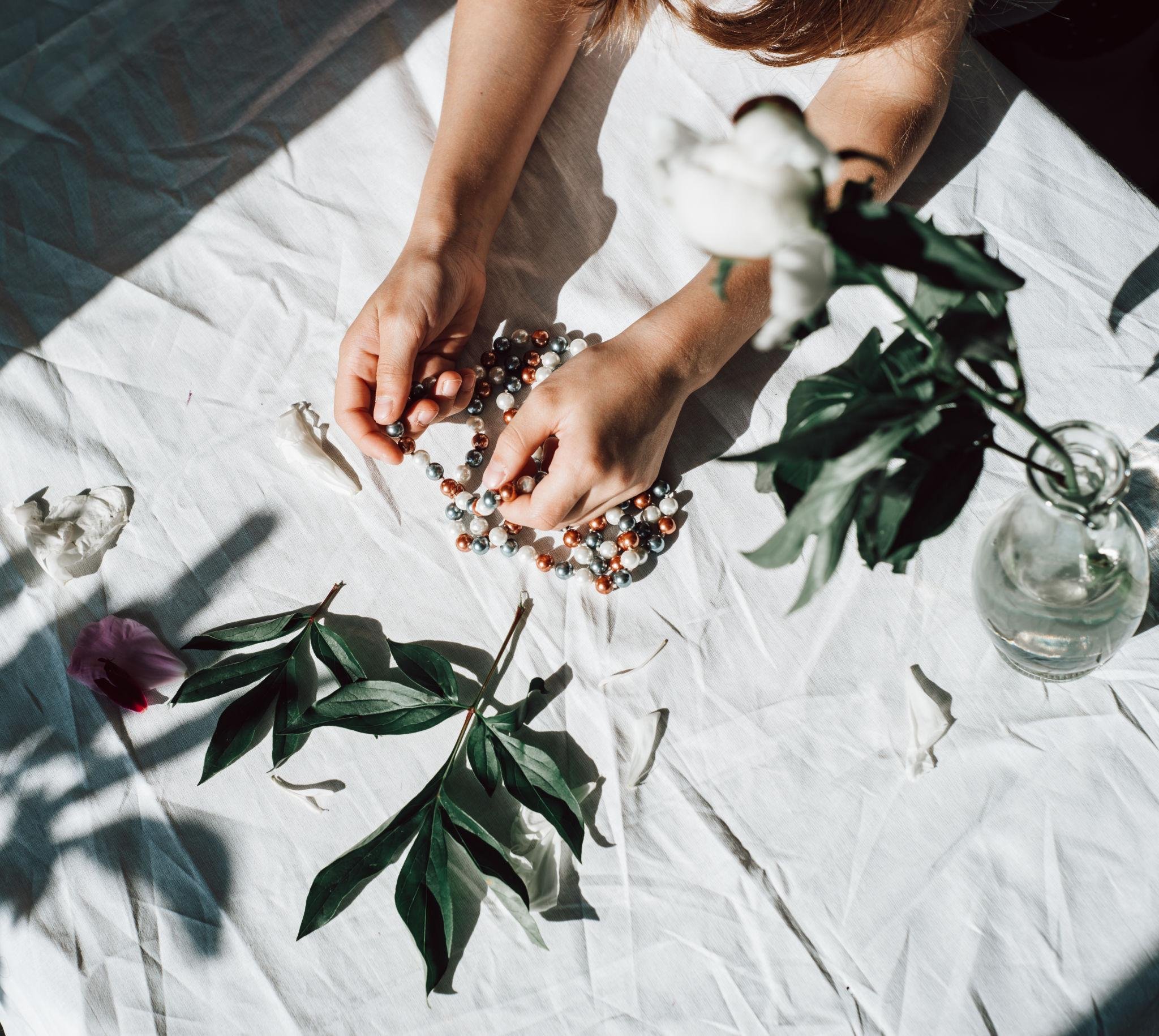 A person arranging a multicolored pearl necklace on a white surface with green leaves and a glass vase with white flowers nearby.