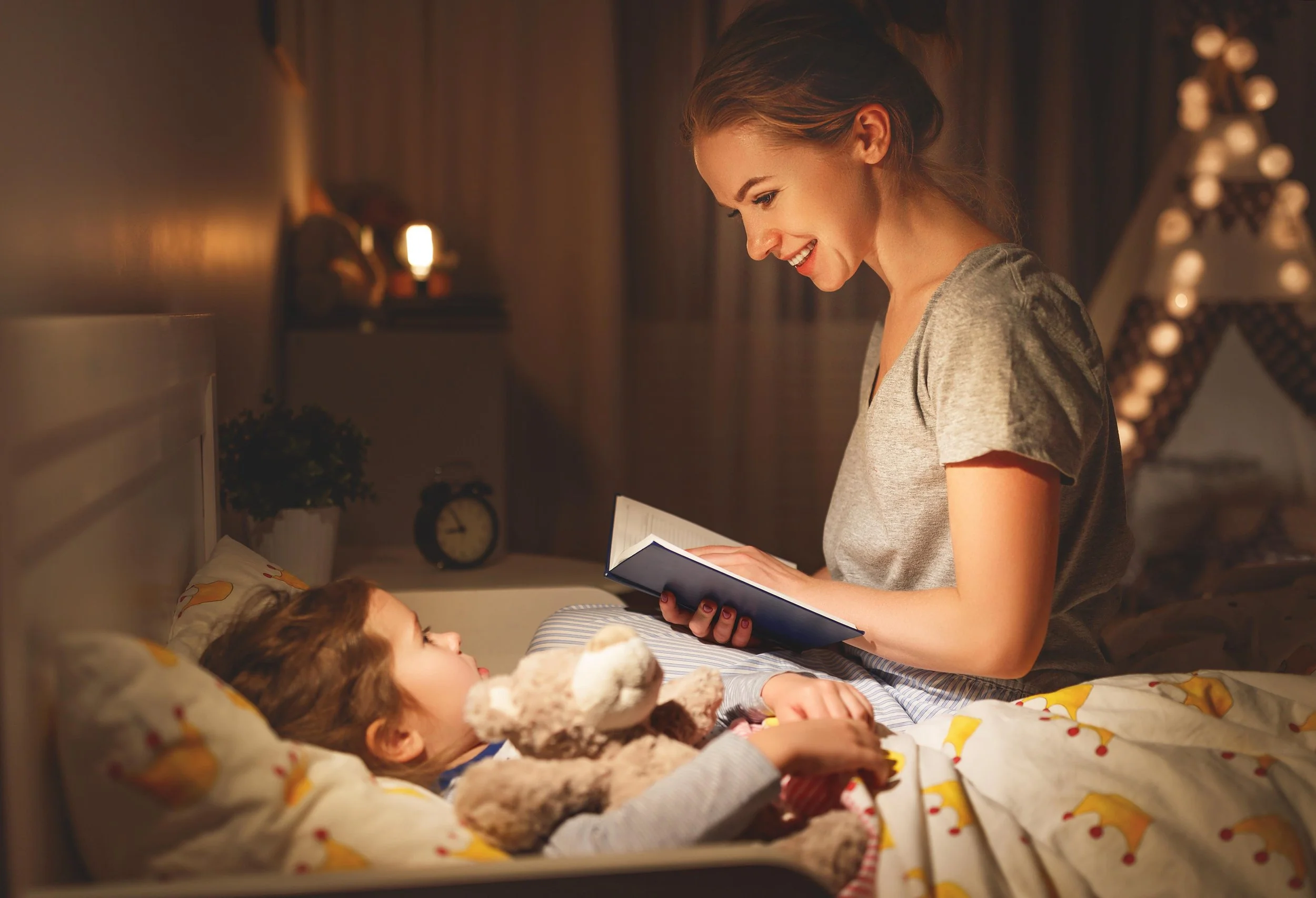 A woman smiles as she reads a book to a young girl lying in bed. The girl looks at the woman and is holding a stuffed animal. The room is warmly lit with a lamp in the background and decorative lights on a Christmas tree.