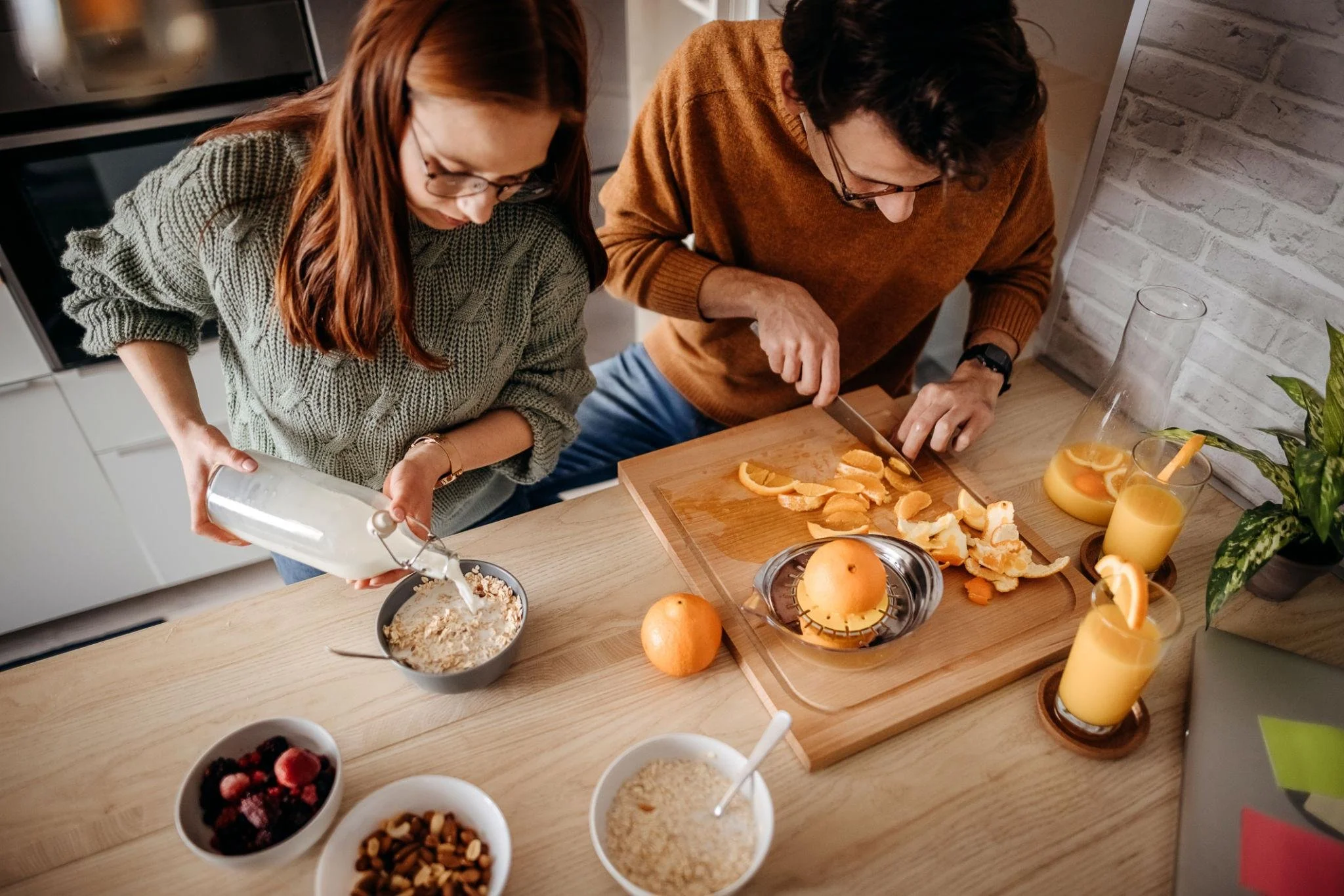 Two people preparing food in a kitchen. One is pouring ingredients from a container into a bowl, and the other is chopping oranges on a wooden cutting board. There are several glasses of orange juice and bowls of berries, nuts, and oats on the table.