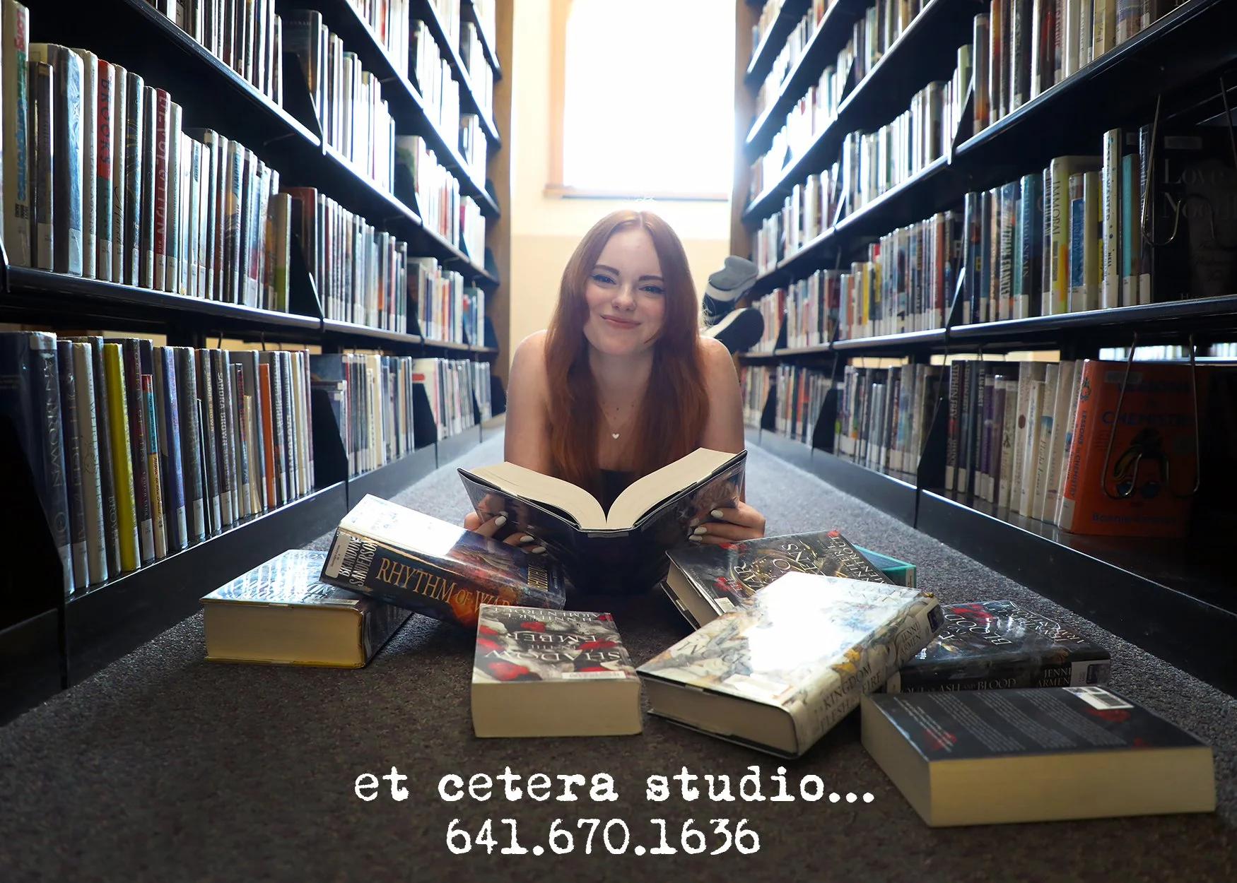 A young woman with long red hair is lying on her stomach on the floor between two bookshelves in a library, surrounded by books. She is holding an open book and has a slight smile. There are several books scattered around her on the floor. The librar
