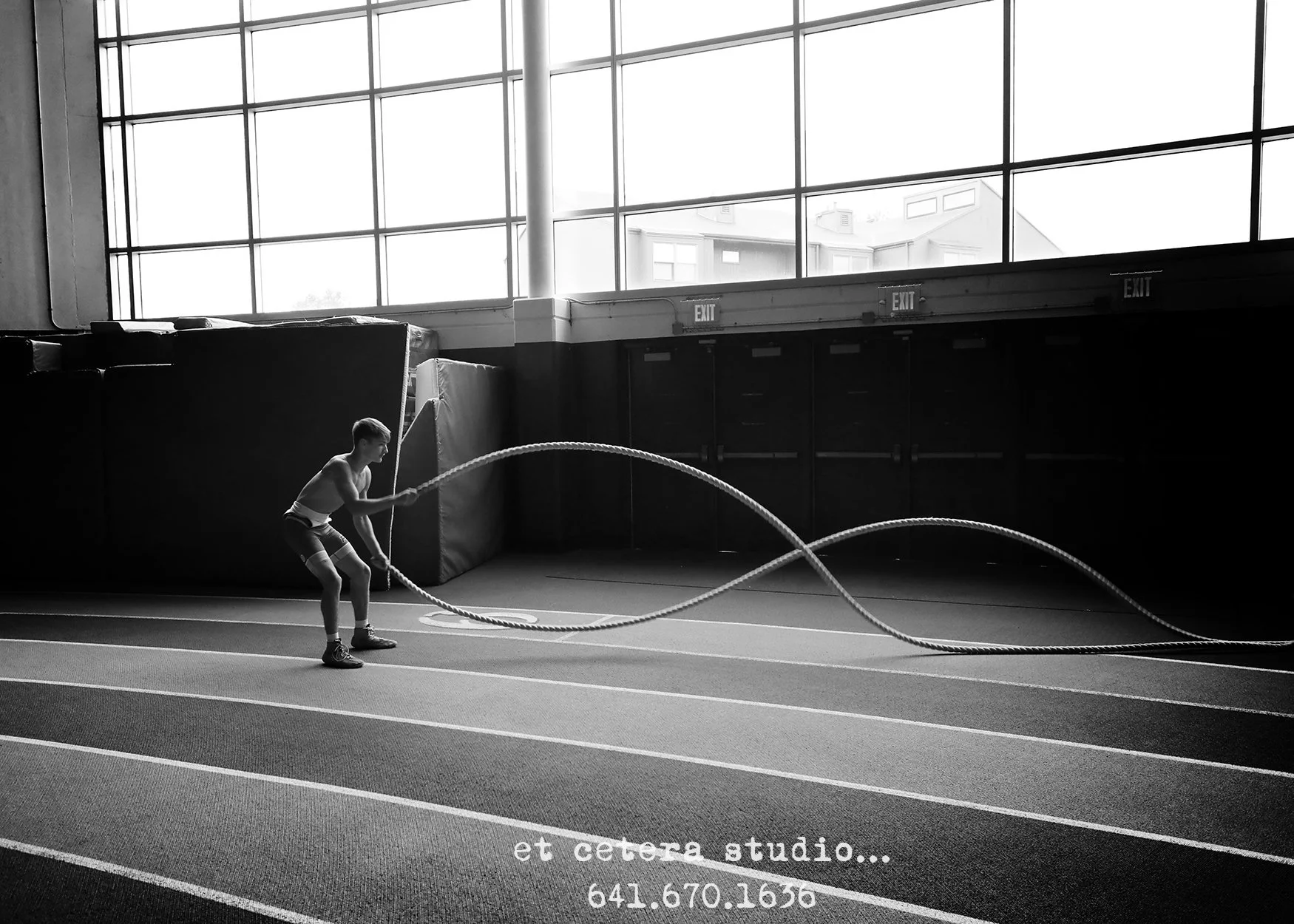 A young boy is exercising with battle ropes inside a gymnasium with large windows, natural light coming in, and visible exit signs.