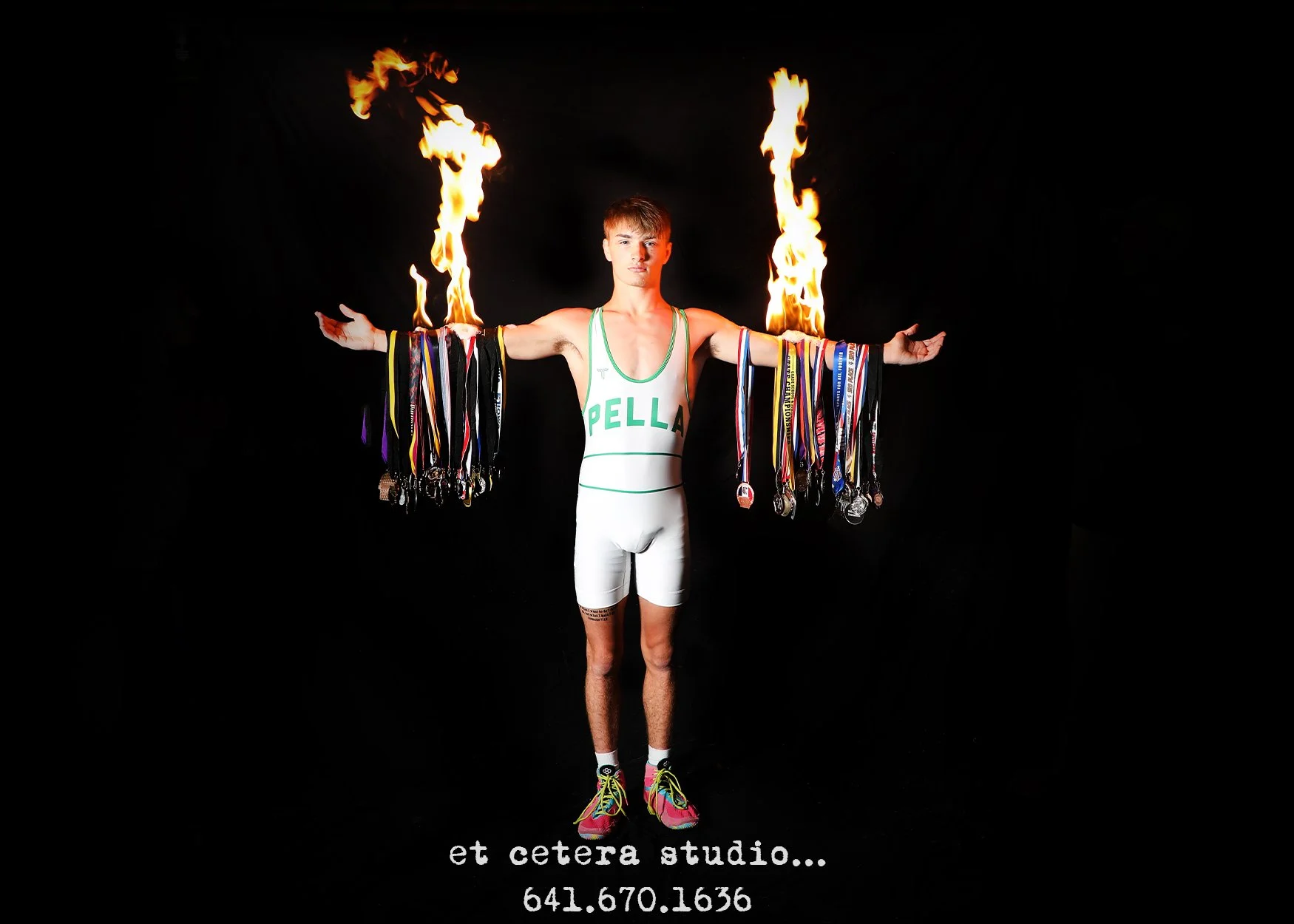 A young male athlete stands with his arms outstretched, holding numerous medals and medals ribbons. He is shirtless, wearing a white athletic uniform with green accents and the word 'PELLA' on the front. Two large flames are burning above each of his