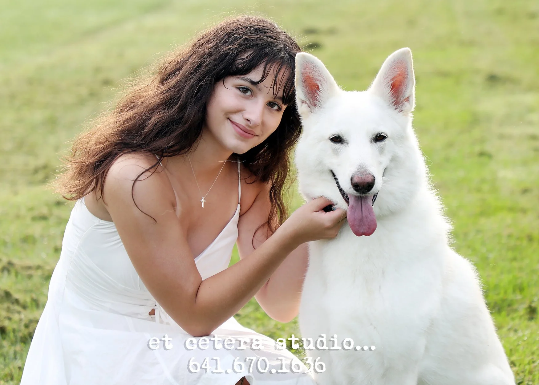 A woman with dark wavy hair wearing a white dress, smiling and petting a large white dog, sitting on grass in an outdoor setting.