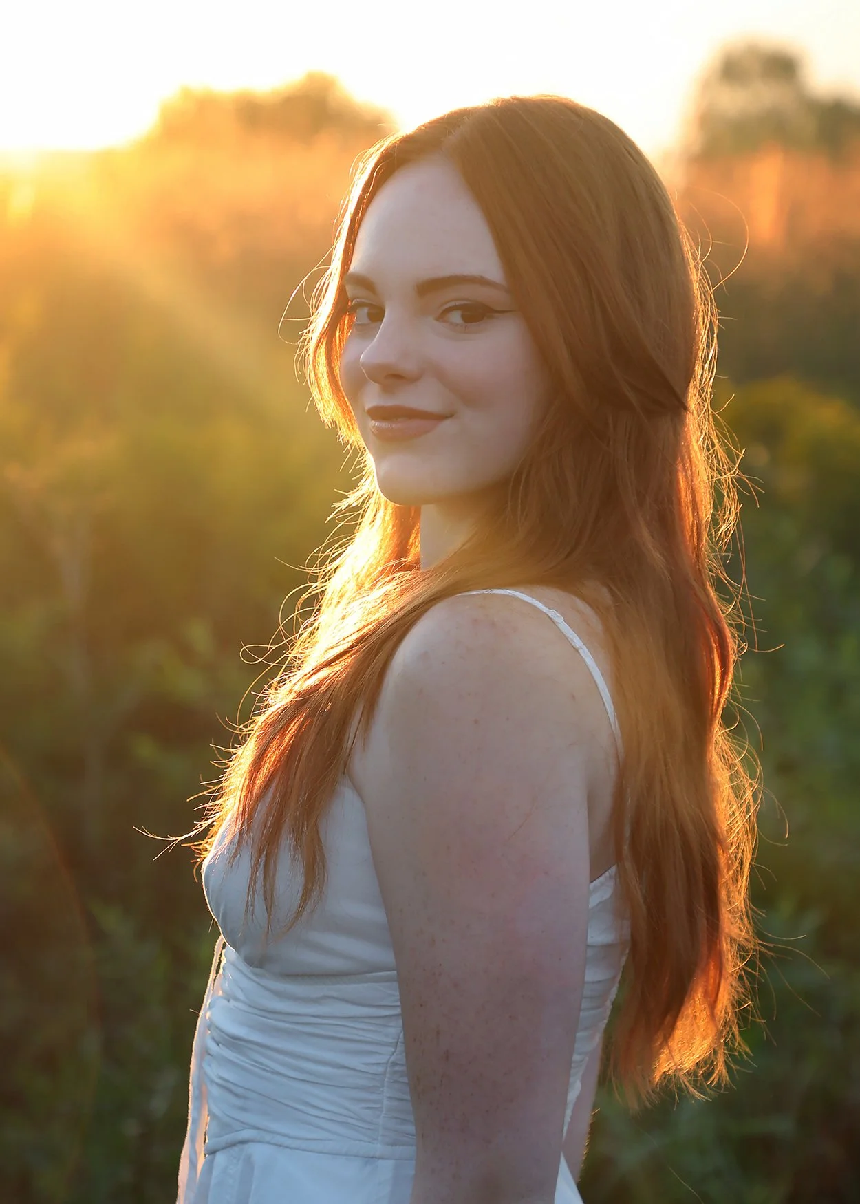 A young woman with long red hair outdoors at sunset, wearing a white dress, looking over her shoulder with a slight smile.