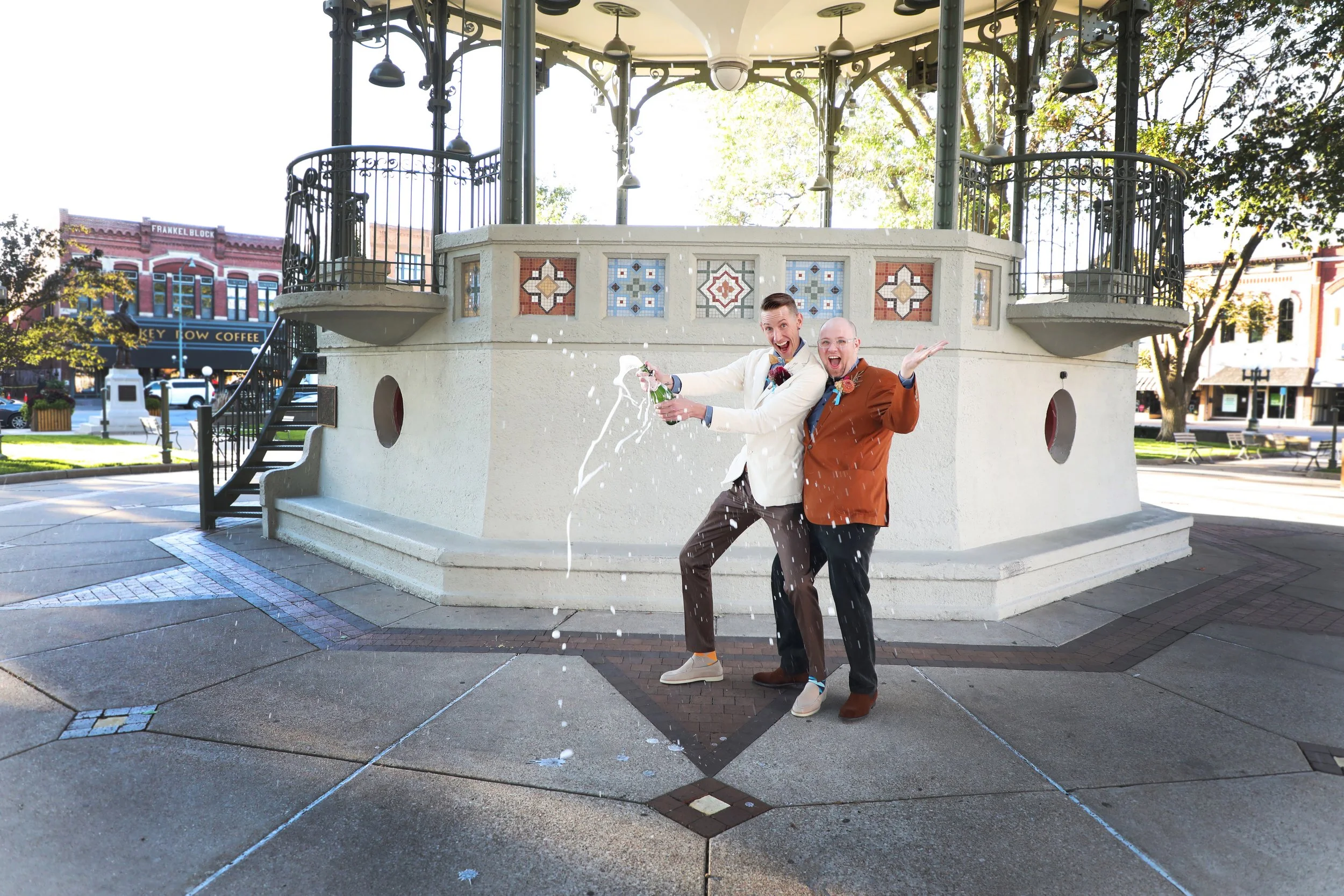 Two men celebrating in front of a gazebo, spraying champagne, in a park with buildings in the background.