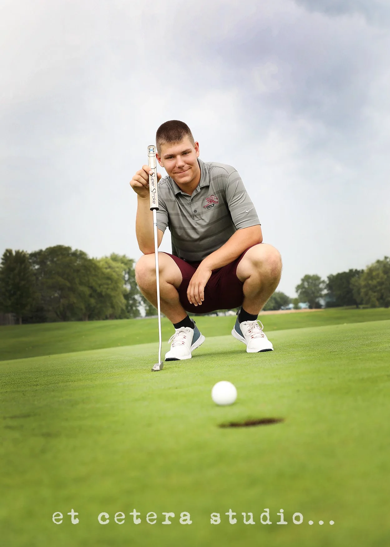 A young man crouching on a golf course, holding a golf club, with a golf ball near the hole, surrounded by green grass and trees in the background.