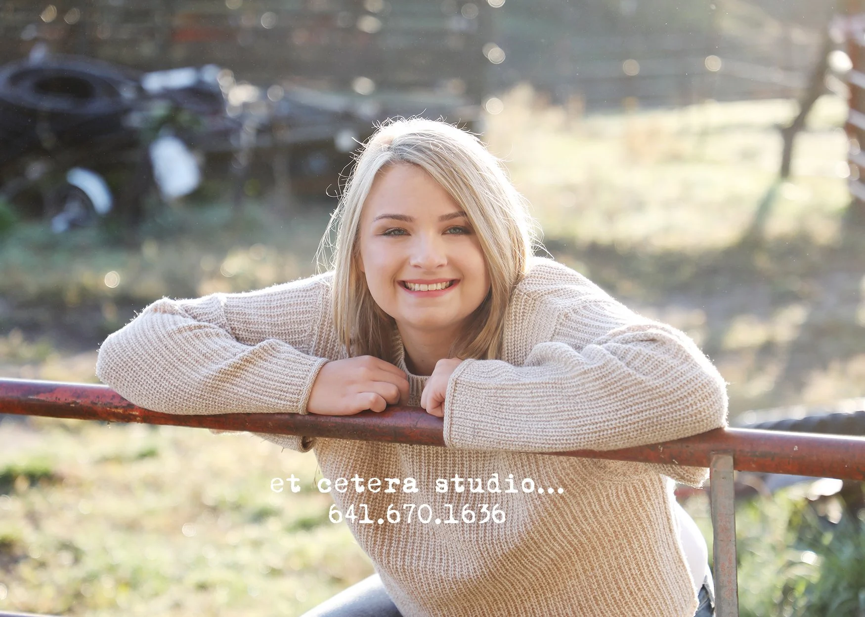 A young woman with blonde hair smiling while resting on a red metal railing outdoors in daylight. Blurred green and brown background with some sunlight.