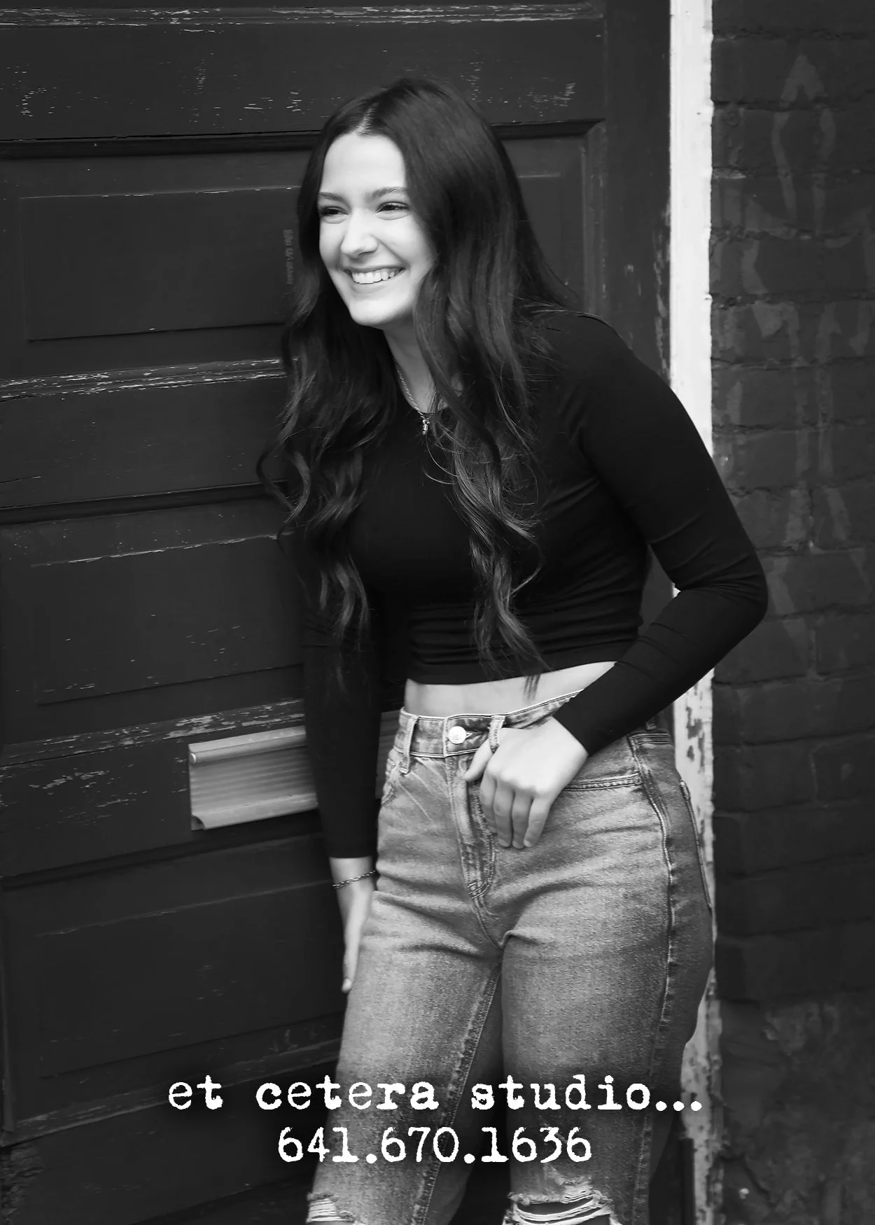 A young woman with long, wavy hair smiling and leaning against a wooden door and brick wall. She is wearing a black long-sleeve crop top and high-waisted jeans. There is text at the bottom promoting a studio with a phone number.
