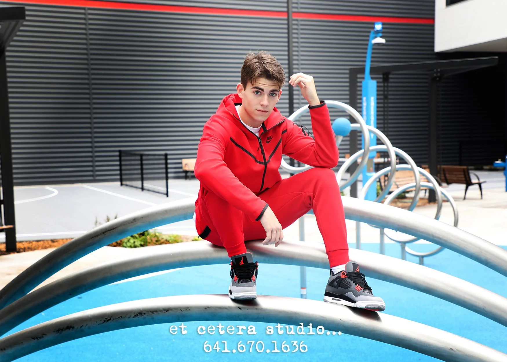 A young man in a red tracksuit, black and red sneakers, and a white shirt, sitting on a curved metal bar at an outdoor playground. He has dark hair, blue eyes, and is looking at the camera with a relaxed pose. The background includes black and red mo