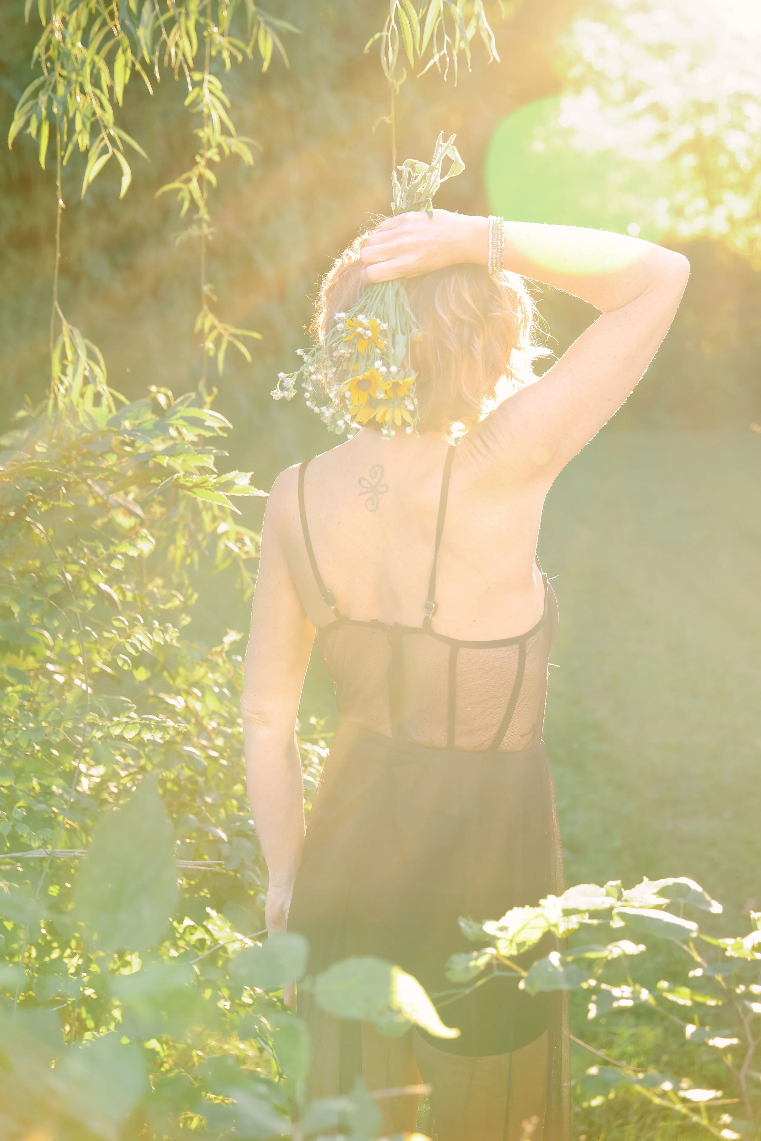 A woman with short hair holding a bouquet of wildflowers behind her head in a sunlit outdoor setting with green foliage and trees.