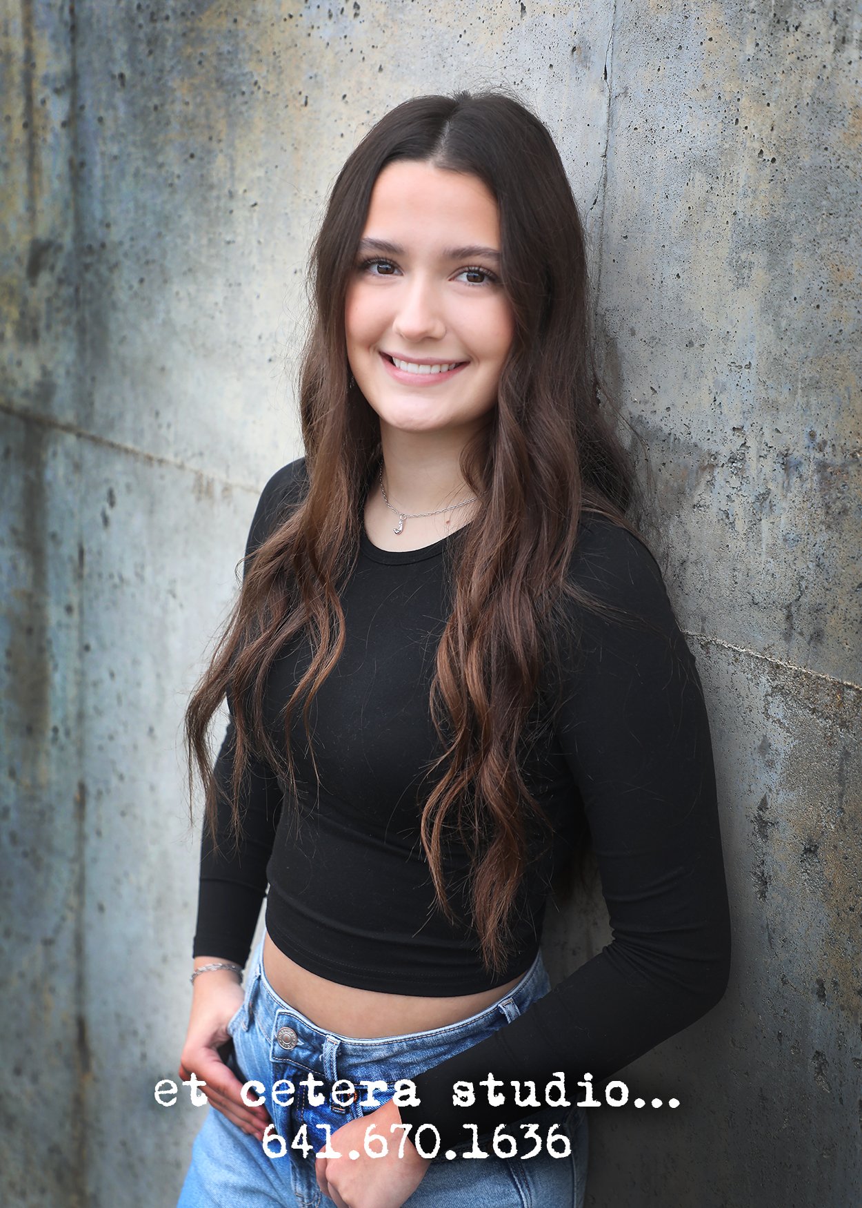 A young woman with long wavy brown hair and light skin, smiling and leaning against a textured concrete wall. She is wearing a black long-sleeve top, blue jeans, and a silver necklace with a small pendant.