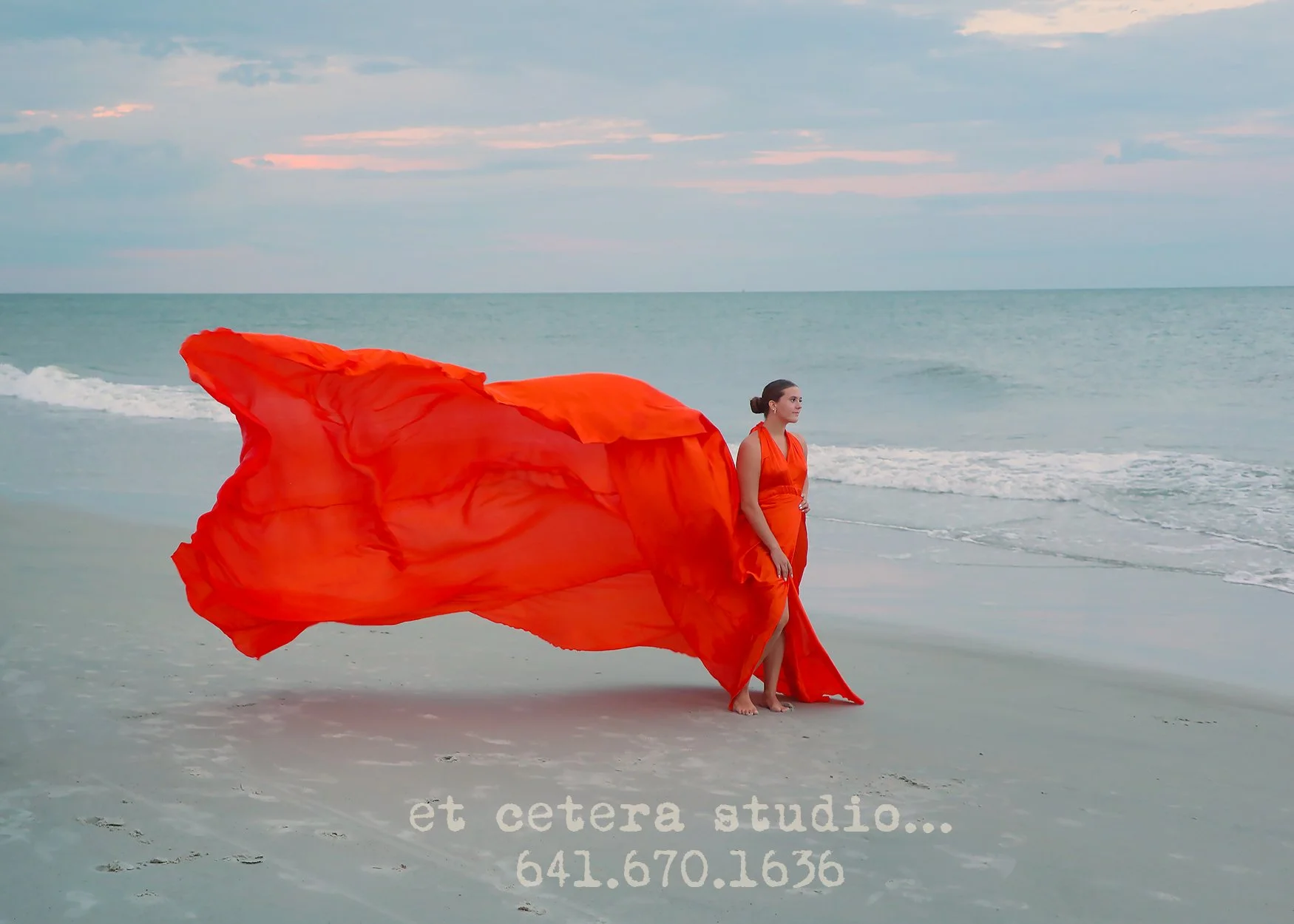 A woman in an orange dress standing on a beach with a large orange fabric flowing behind her, near the ocean, with cloudy sky above.