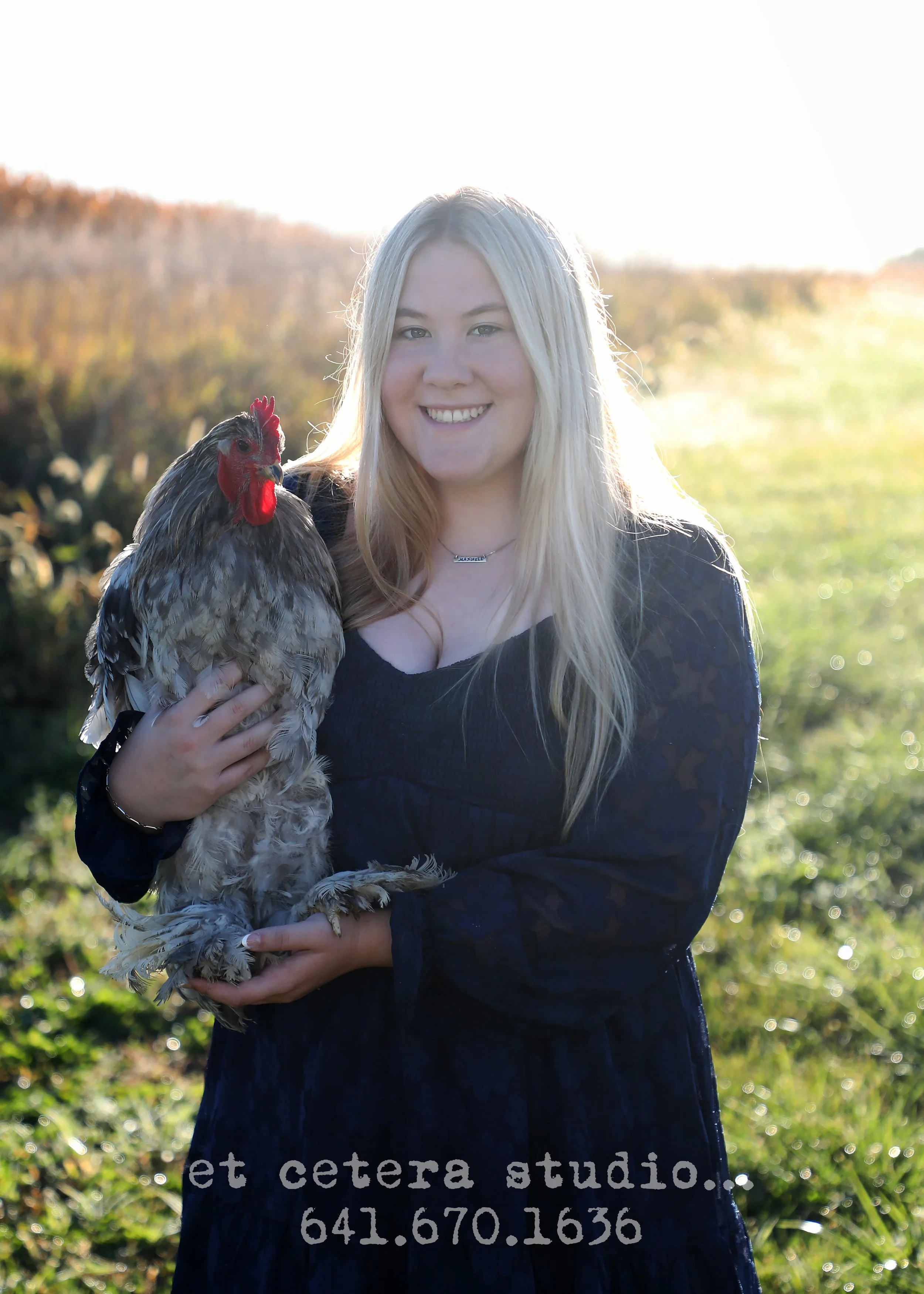 A woman with long blonde hair smiling outdoors while holding a chicken in her arms, with a grassy field and trees in the background during daylight.