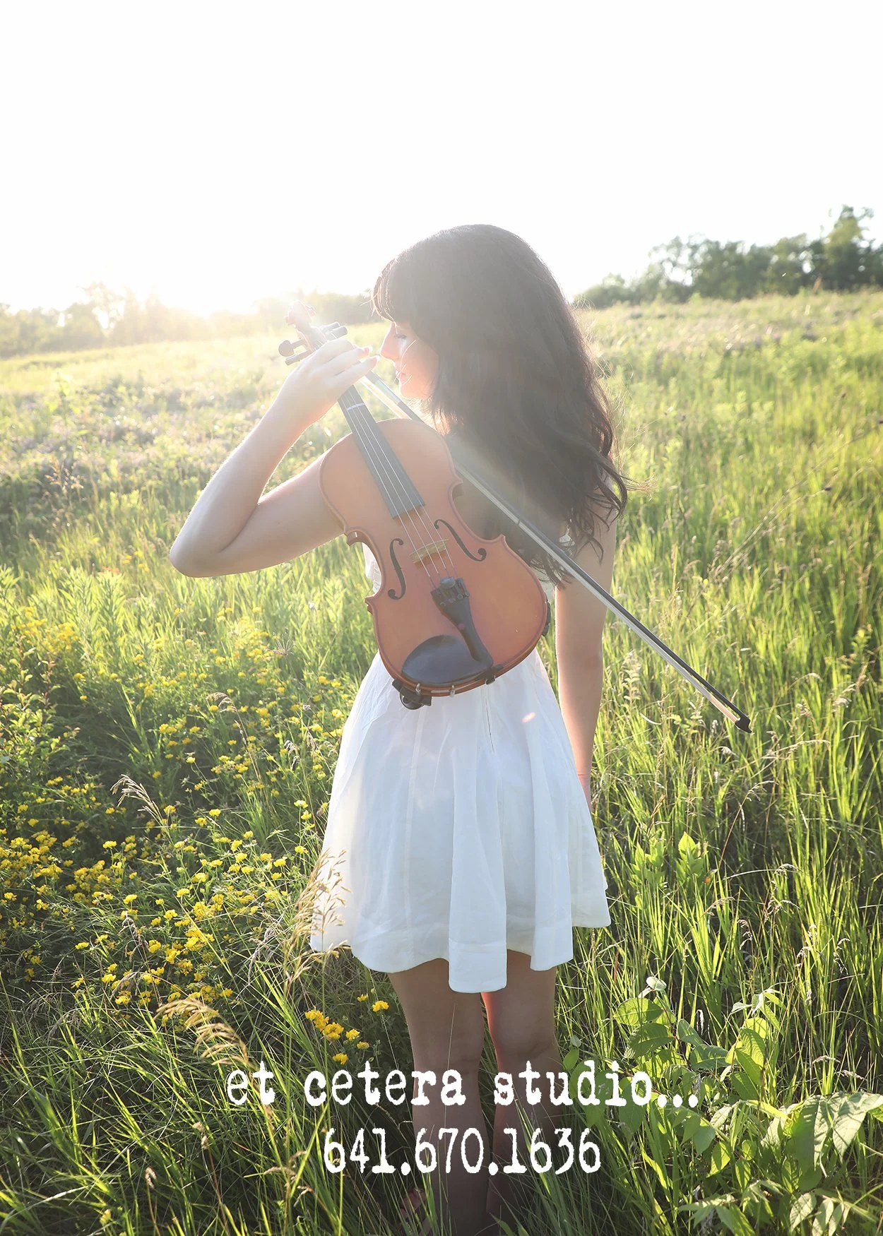A woman in a white dress playing a violin in a grassy field during sunset, with sunlight creating a glow around her.