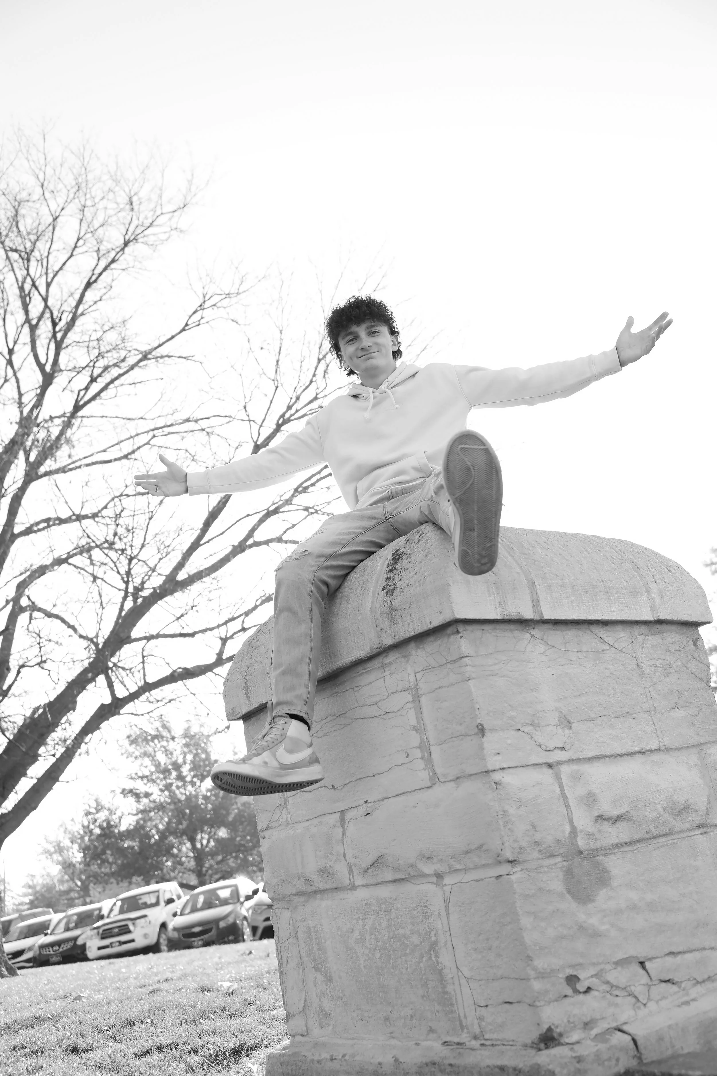A young man with curly hair is sitting on a large stone monument with his arms outstretched, smiling slightly. Bare tree branches are visible in the background, and a row of parked cars is seen at the bottom of the image.
