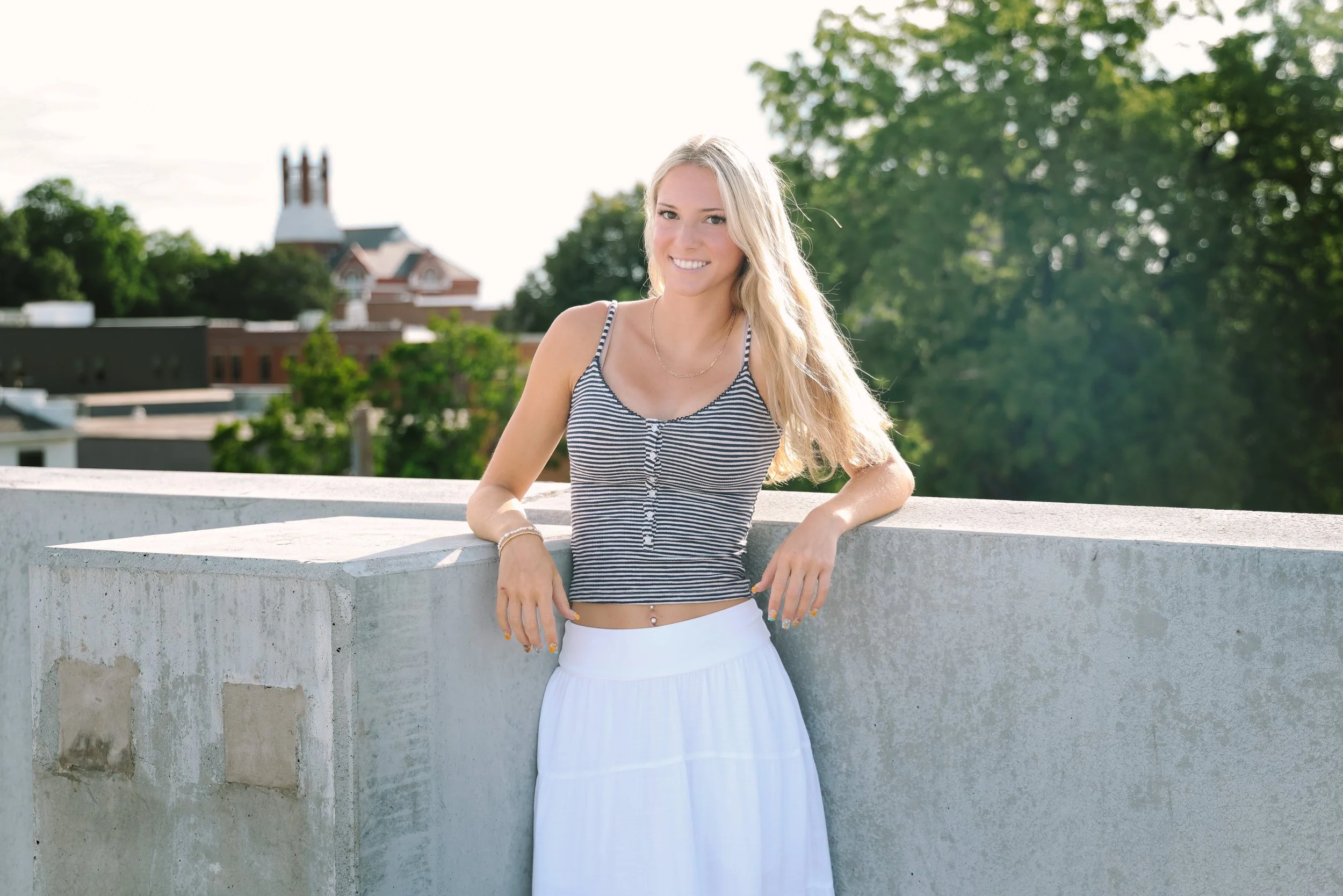 A young woman with long blonde hair smiling, standing outdoors near a concrete wall with trees and a building with a clock tower in the background.