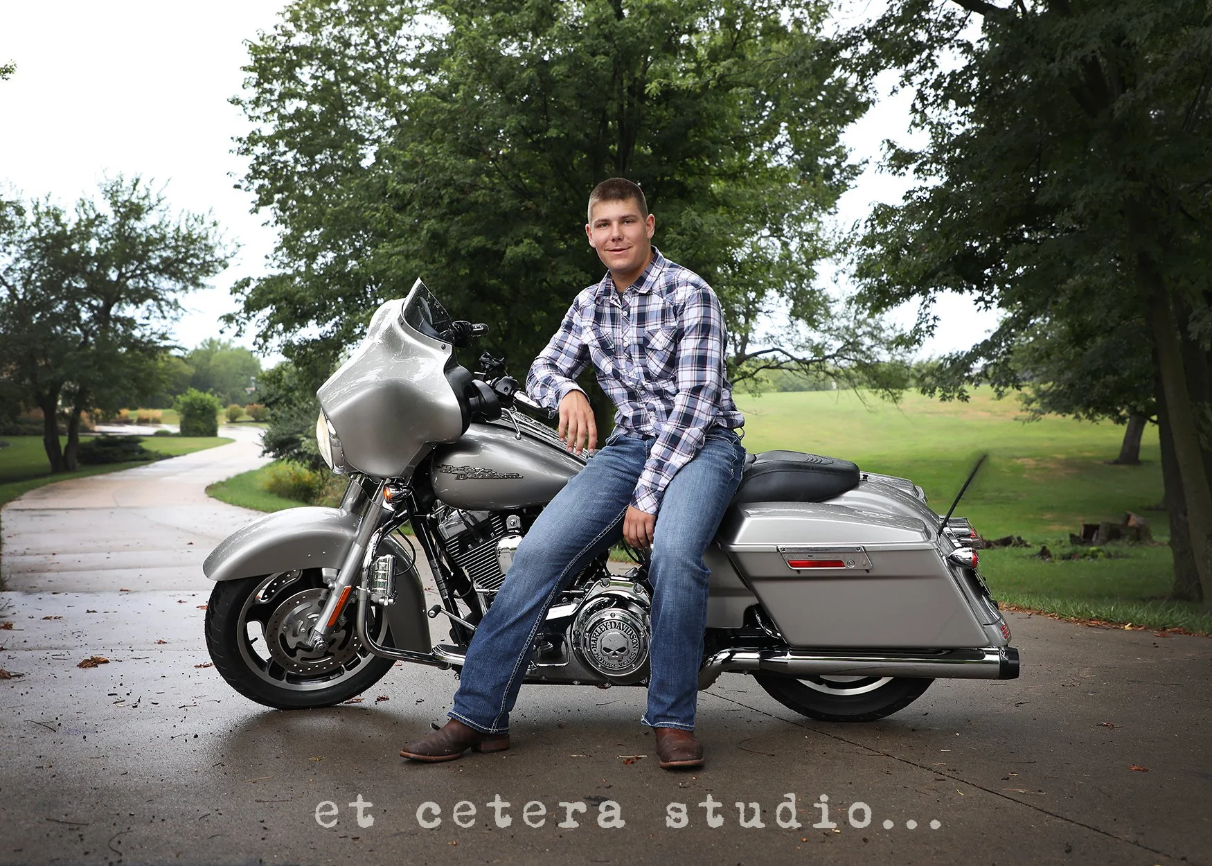 A young man in a plaid shirt and jeans sitting on a silver Harley Davidson motorcycle on a paved road in a park with trees and grass in the background.