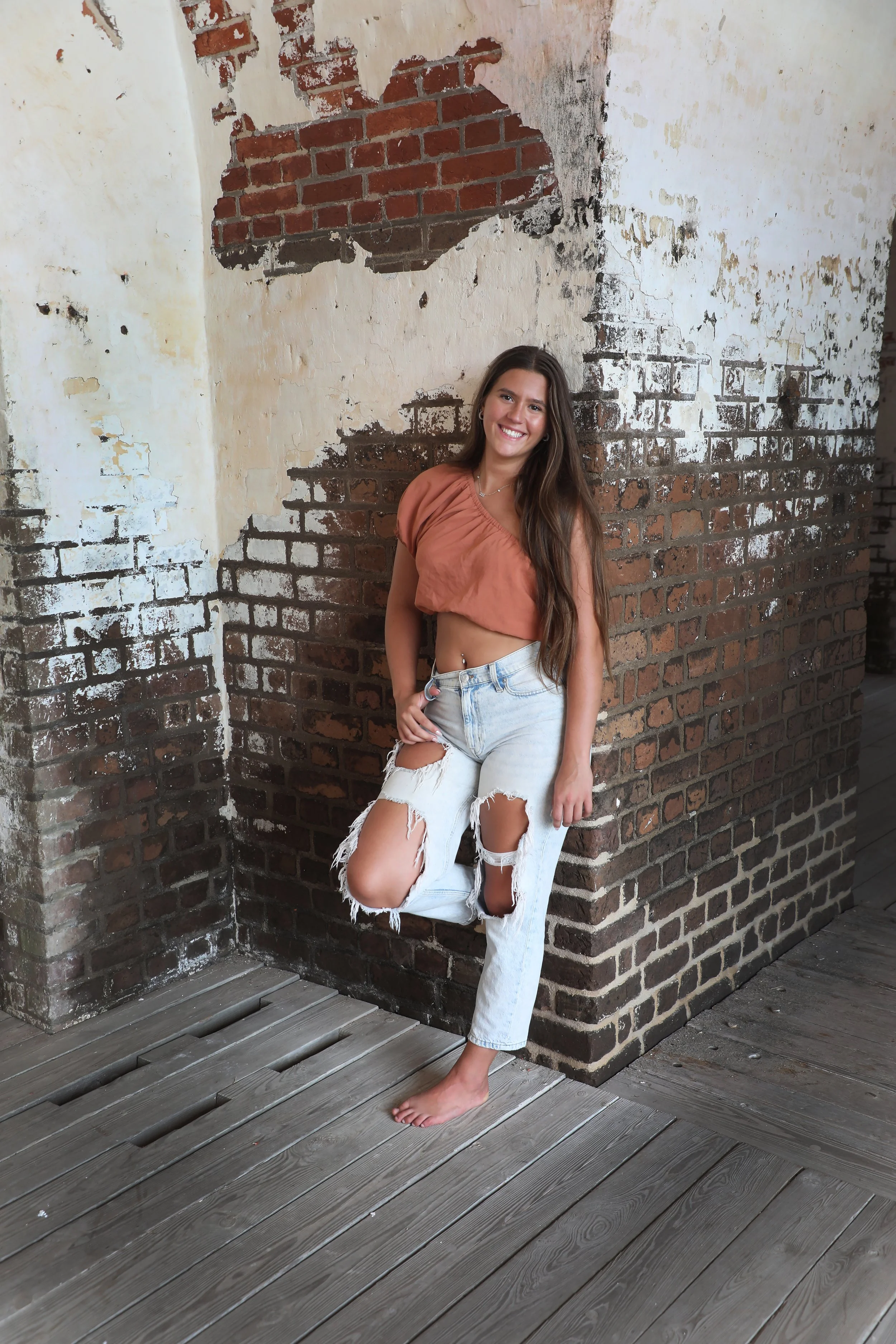 A young woman with long brown hair smiling, wearing a brown crop top and ripped white jeans, standing barefoot against a weathered brick wall with peeling paint, on a wooden deck.