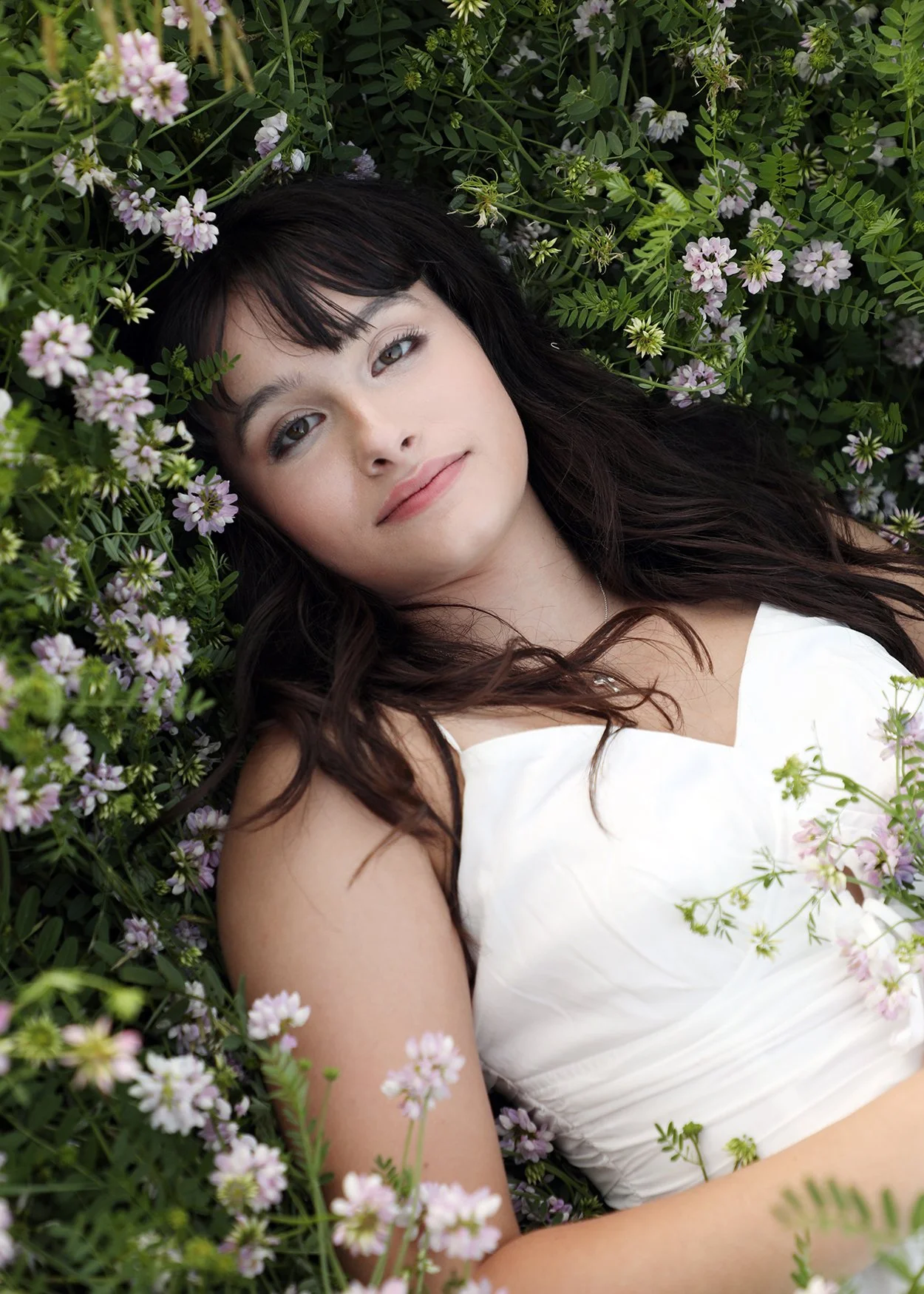 A woman with long dark hair lying among green foliage and pink flowers, wearing a white dress, gazing softly at the camera.