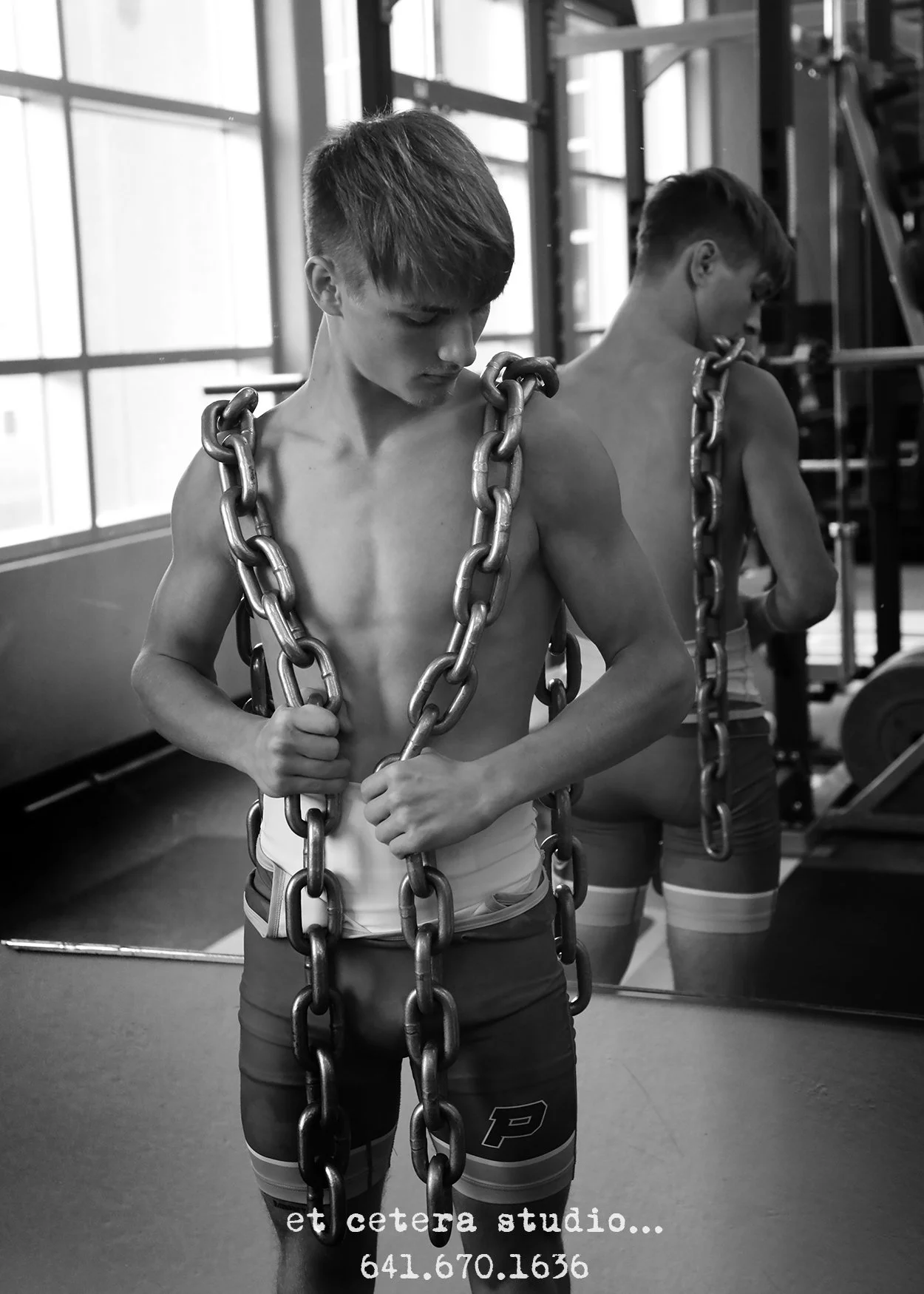 A shirtless young man in workout shorts with a chain necklace stands in front of a mirror in a gym. He appears to be focused on his reflection, with the background showing gym equipment and large windows.