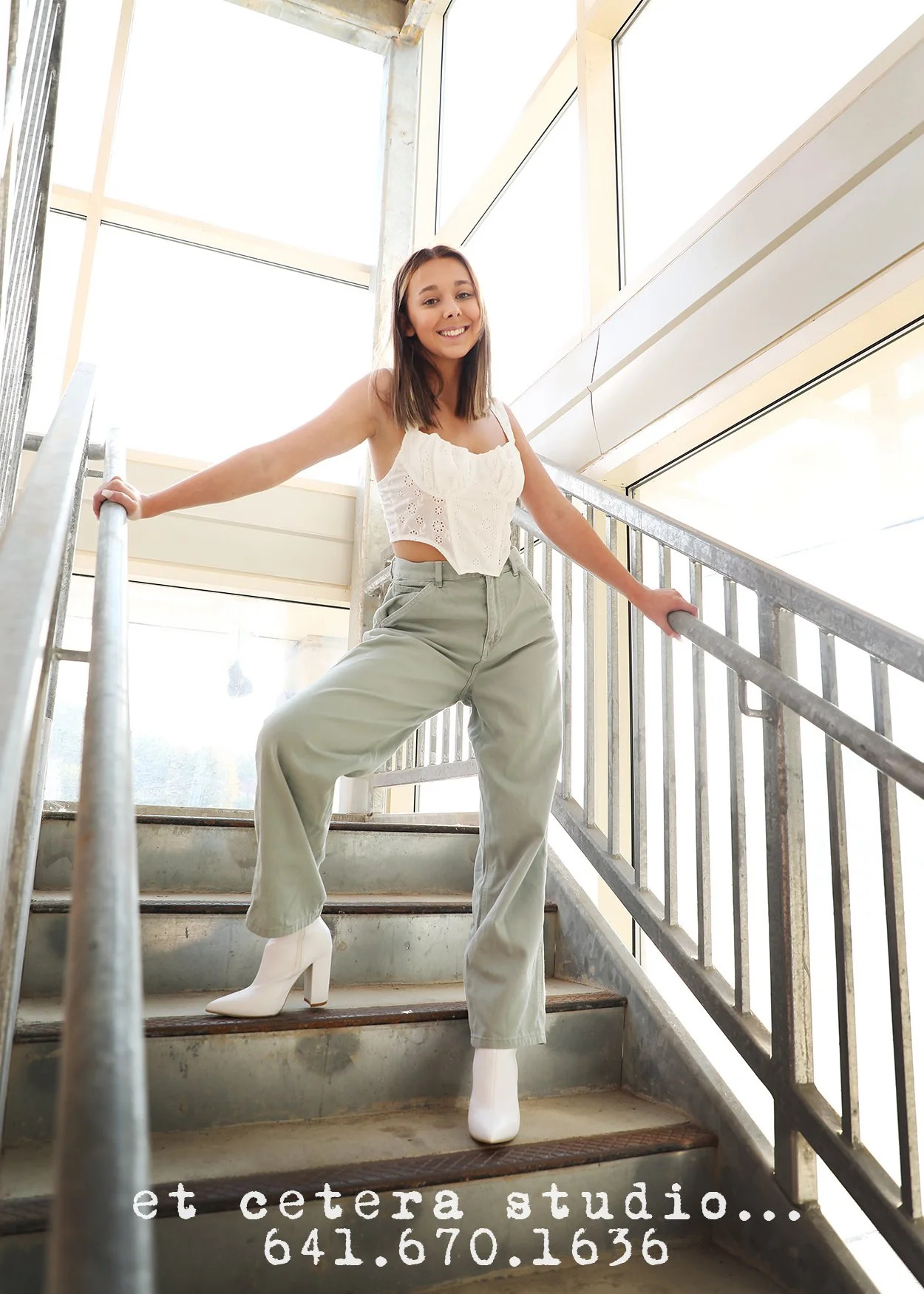 Young woman standing on staircase in bright indoor space, smiling, wearing a white eyelet top, light-colored high-waisted pants, white ankle boots, with a metal railing on her sides, and large windows above. Text overlay with studio name and phone nu