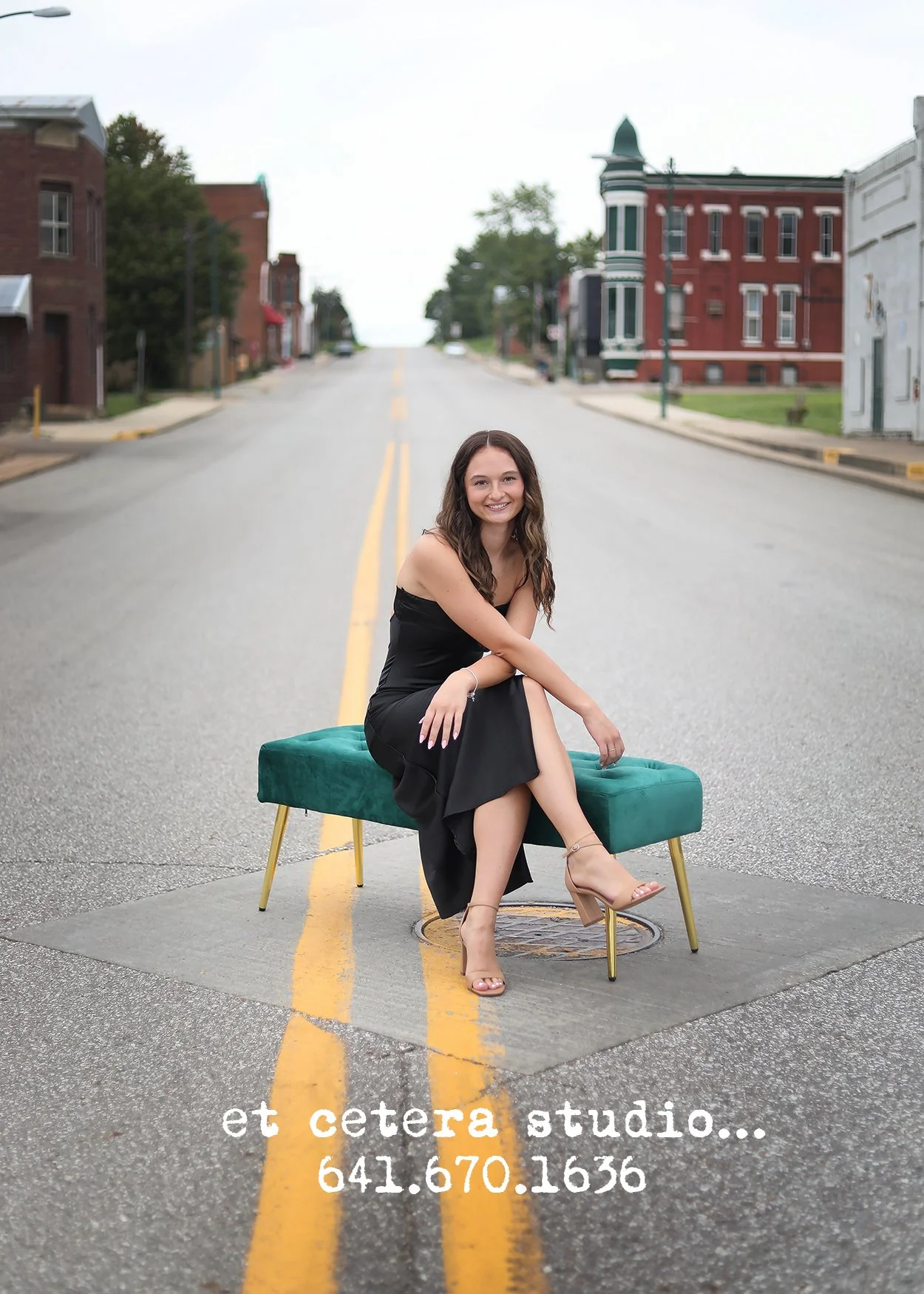 A woman in a black dress and high heels sitting on a teal bench in the middle of a street with a slight crosswalk, smiling at the camera. The street is in a small town with brick buildings on both sides. Text overlay reads "et cetera estudio... 641.6
