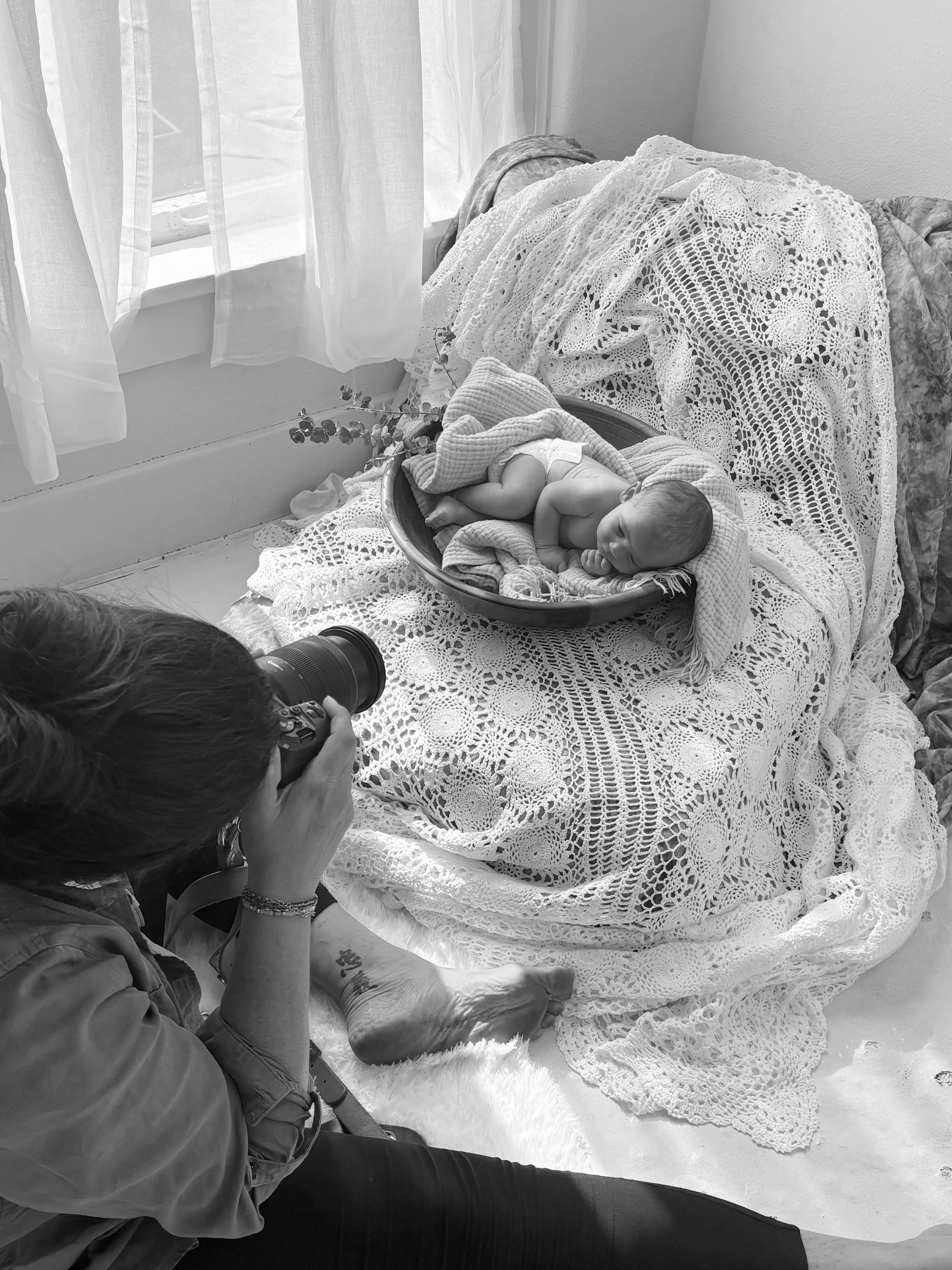 A woman taking a photograph of a newborn baby lying in an oval basket, surrounded by a knitted blanket, with a lace cover in a sunlit room.