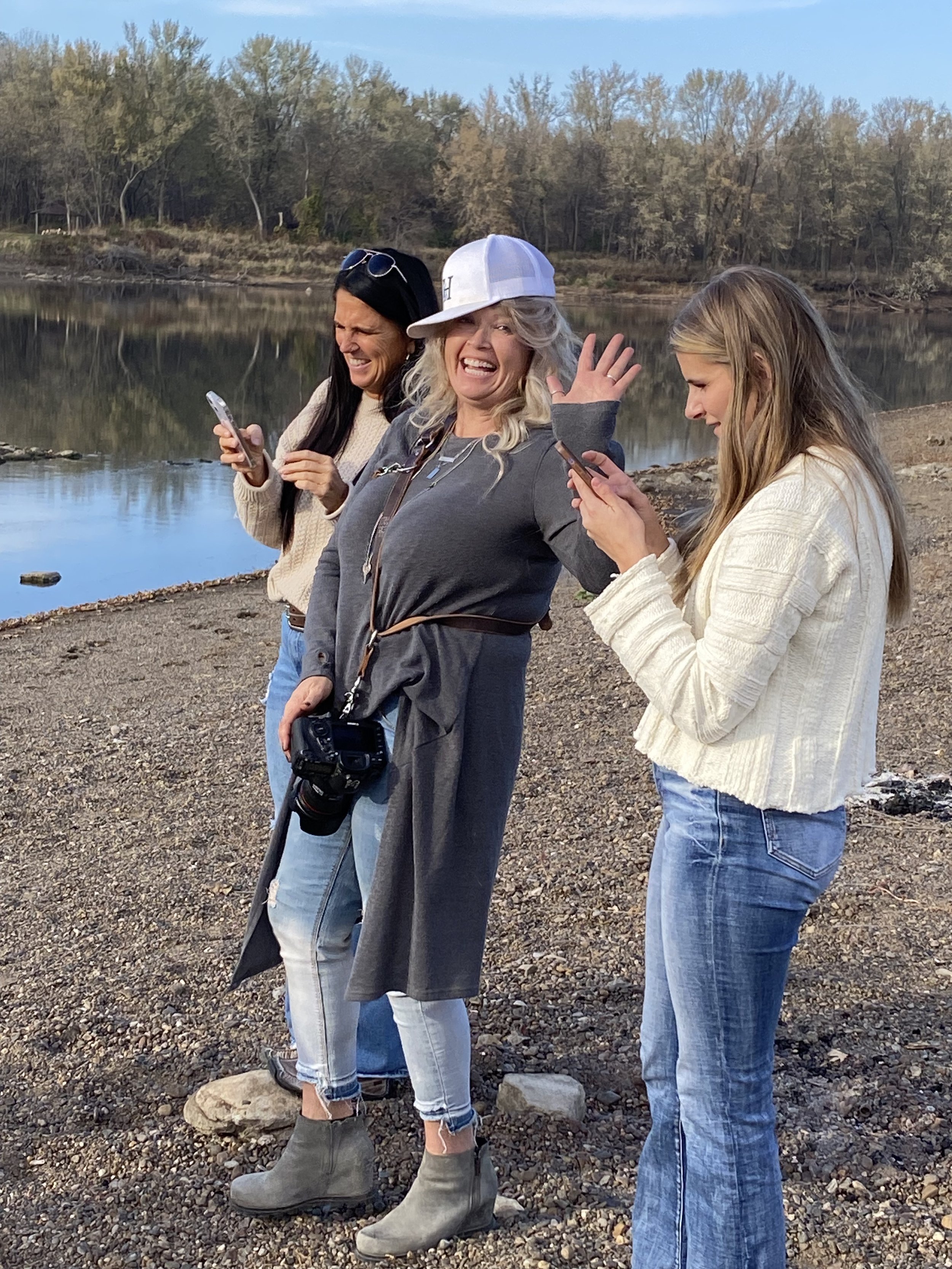 Three women standing on a rocky shoreline by a body of water, looking at their phones and smiling. One woman is waving. They are dressed casually with trees in the background.