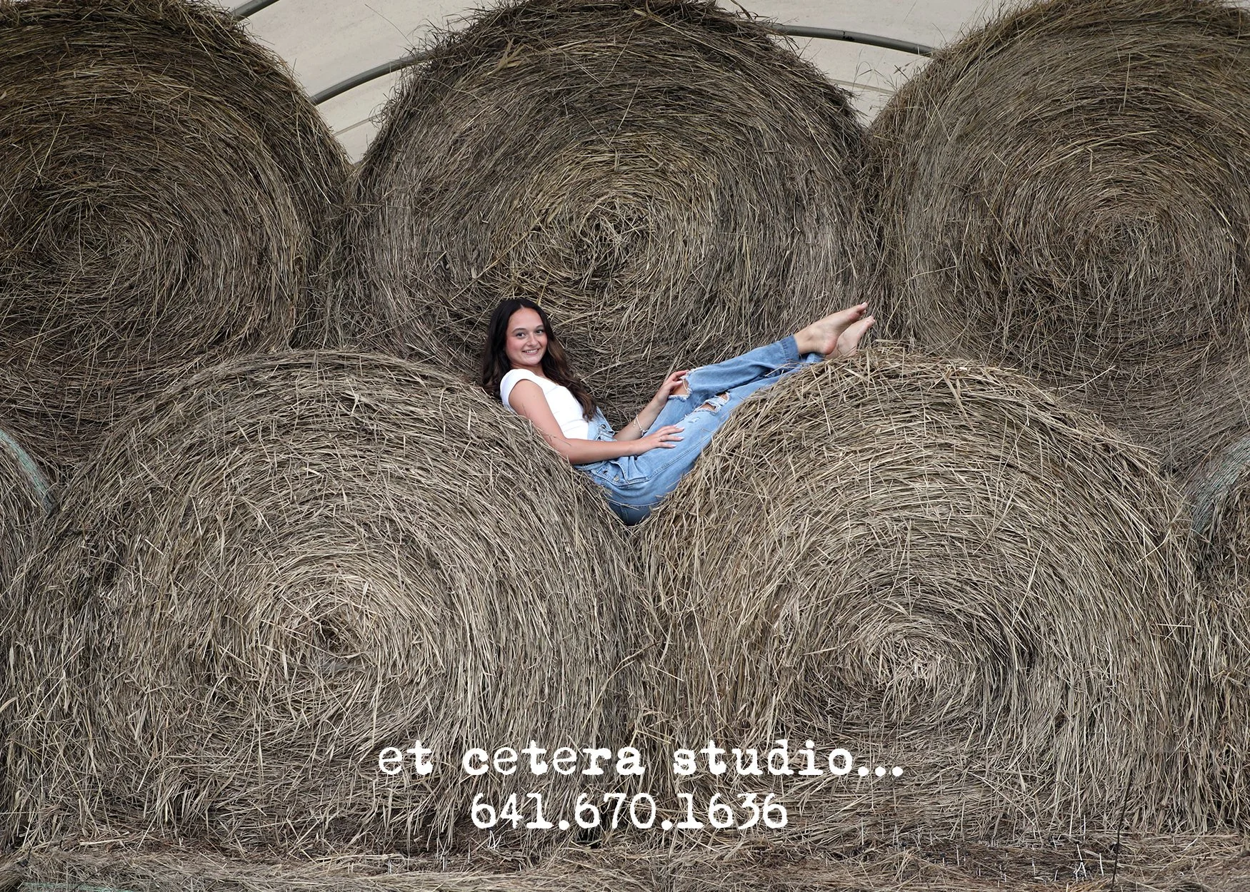 A young woman with long dark hair, wearing a white t-shirt and ripped jeans, smiling while reclining on a large hay bale in a barn or farm setting.