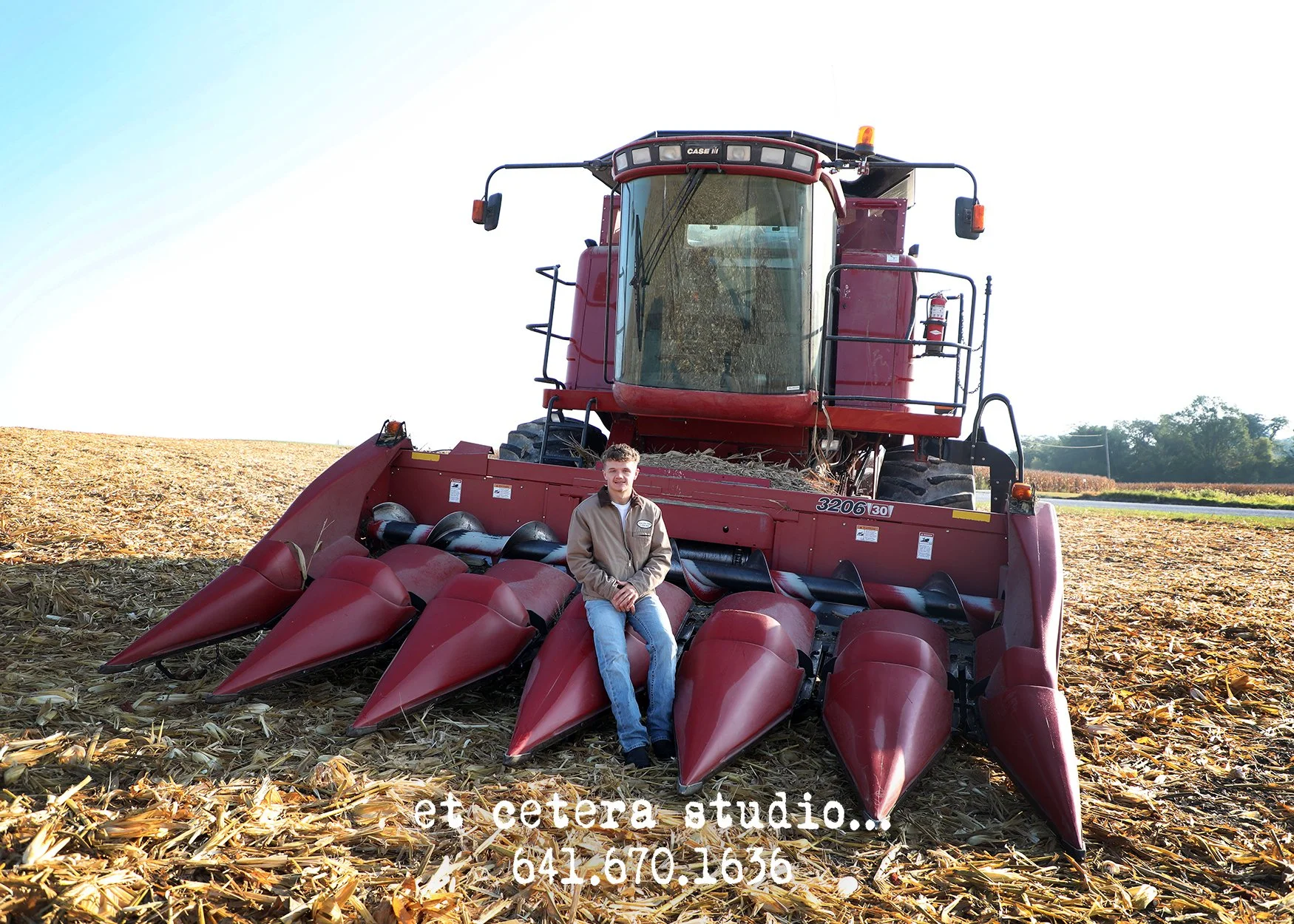 A person sitting on a large red combine harvester in a harvested cornfield on a sunny day with a clear sky and trees in the background.
