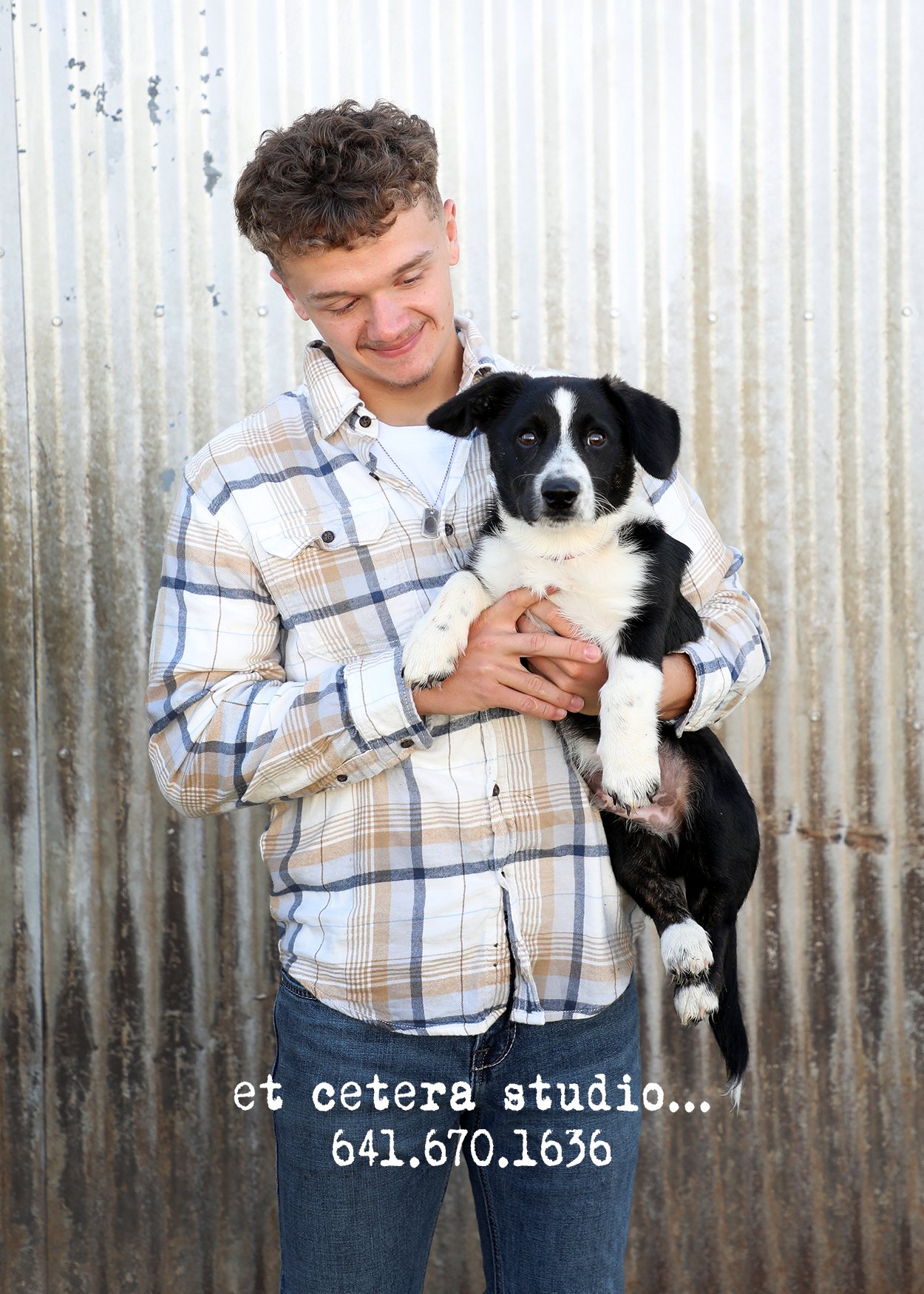 A smiling young man in a plaid shirt holding a black and white puppy in front of a corrugated metal wall. Text overlay: "et cetera studio... 641.670.1636".