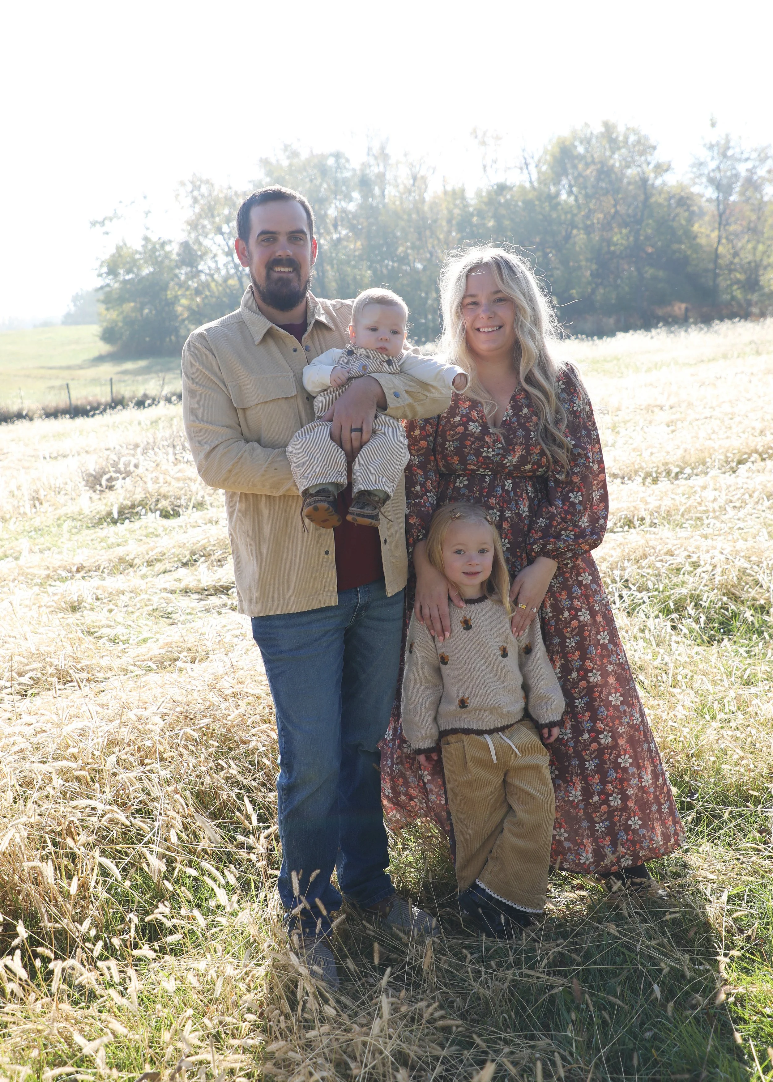 A family of five standing outdoors in a field with trees in the background, enjoying a sunny day.